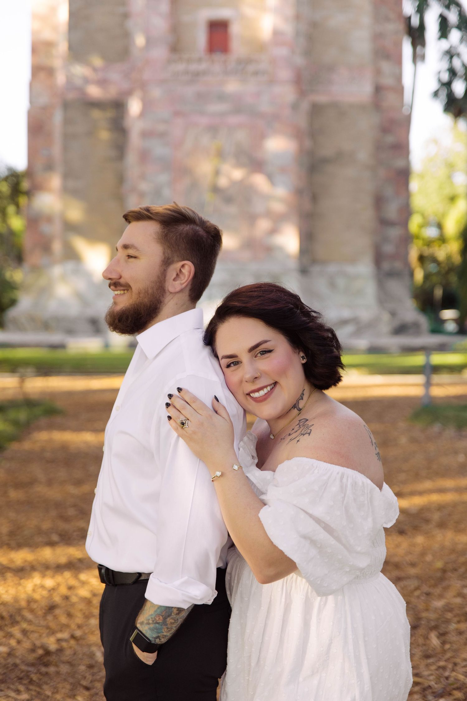 Smiling bride in white off-shoulder dress embraces bearded groom outdoors near stone tower at Bok Tower Gardens, Lake Wales.