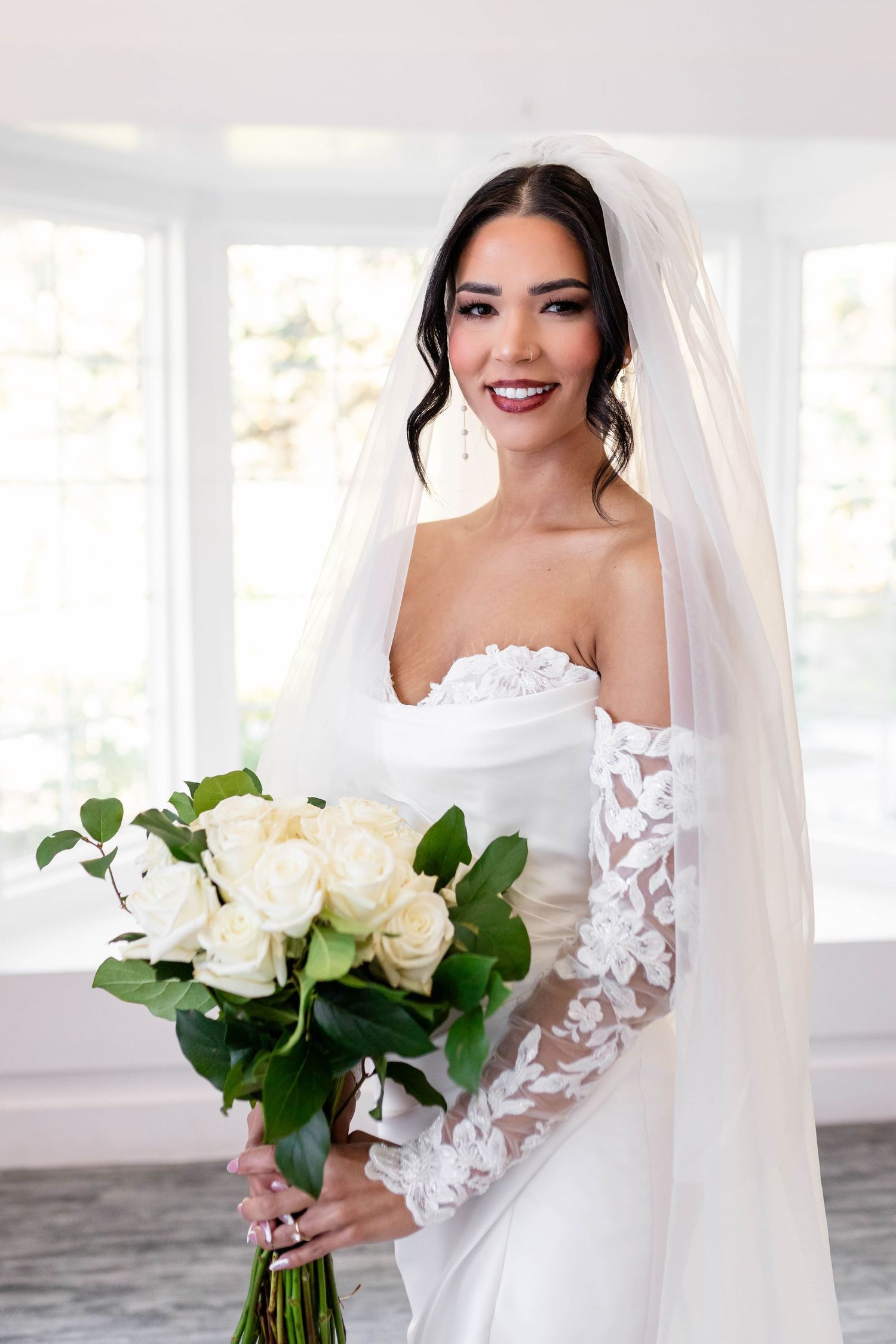 Stunning bride in off-shoulder lace gown and veil holds a bouquet of white roses.