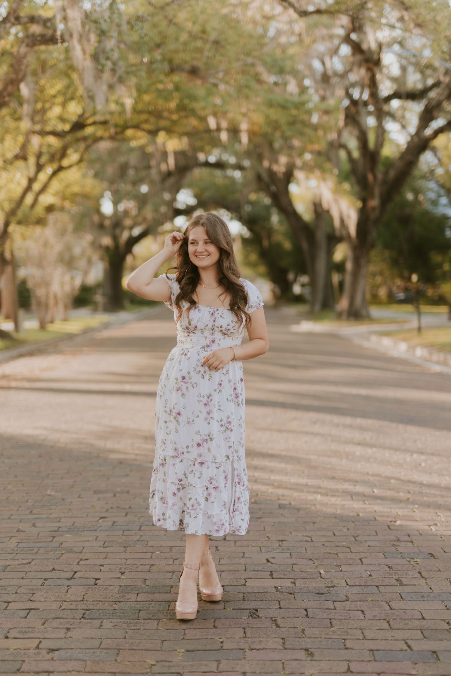 Young woman in floral midi dress and heels posing on brick path lined with moss-draped trees.