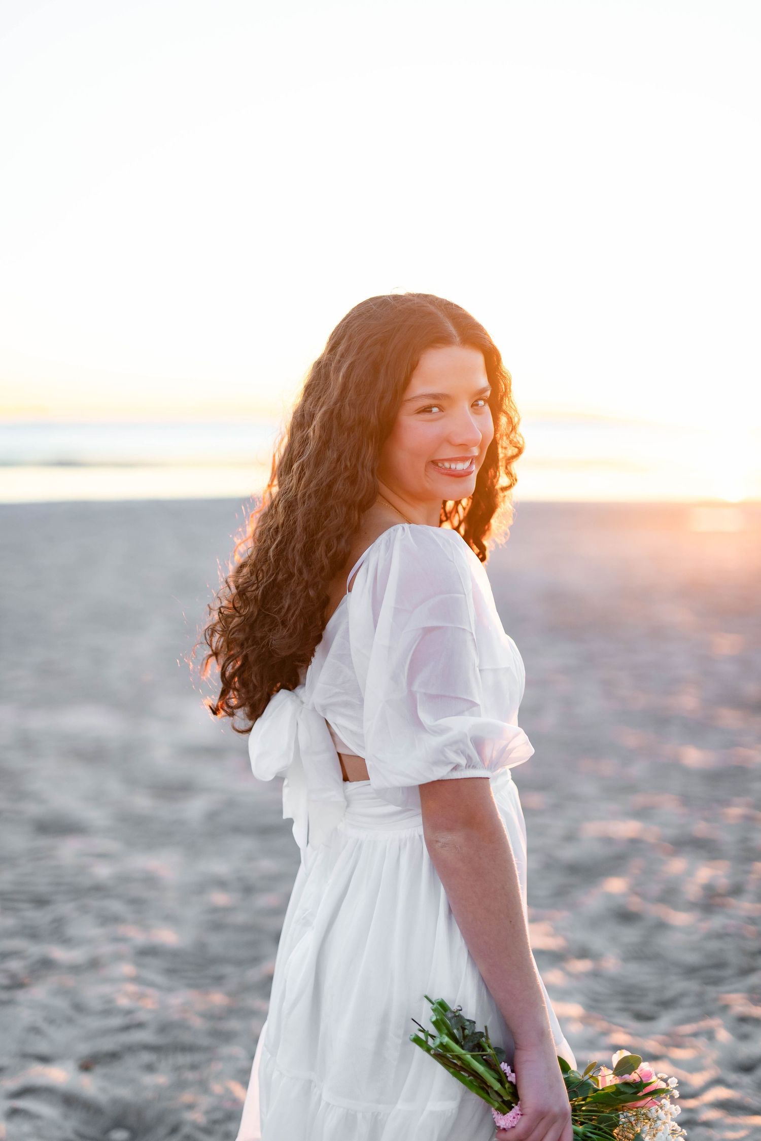 Smiling woman in white dress holding flowers on beach at sunset with curly hair blowing in the breeze.