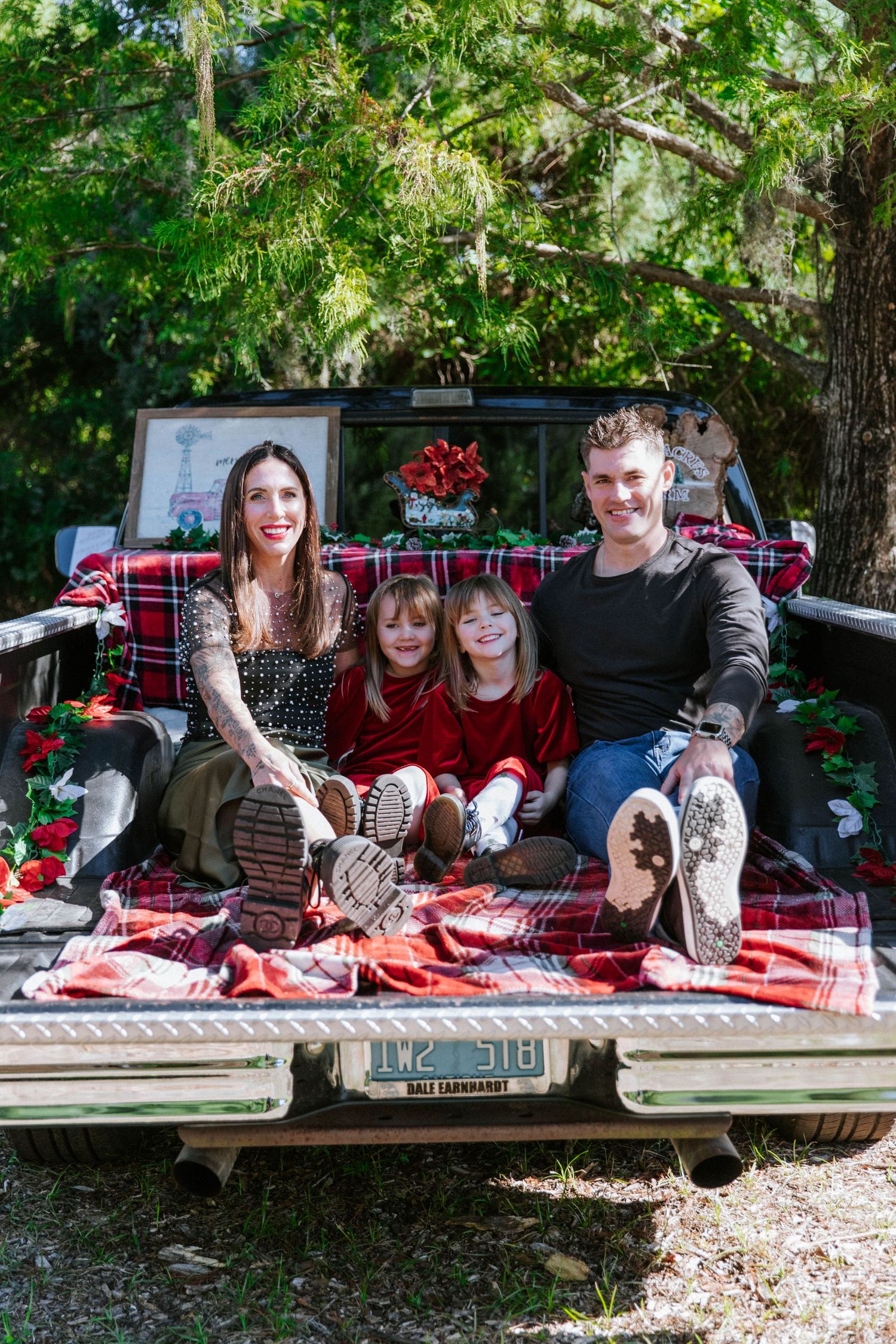 Family of four sitting in truck bed with plaid blanket and Christmas decorations outdoors.