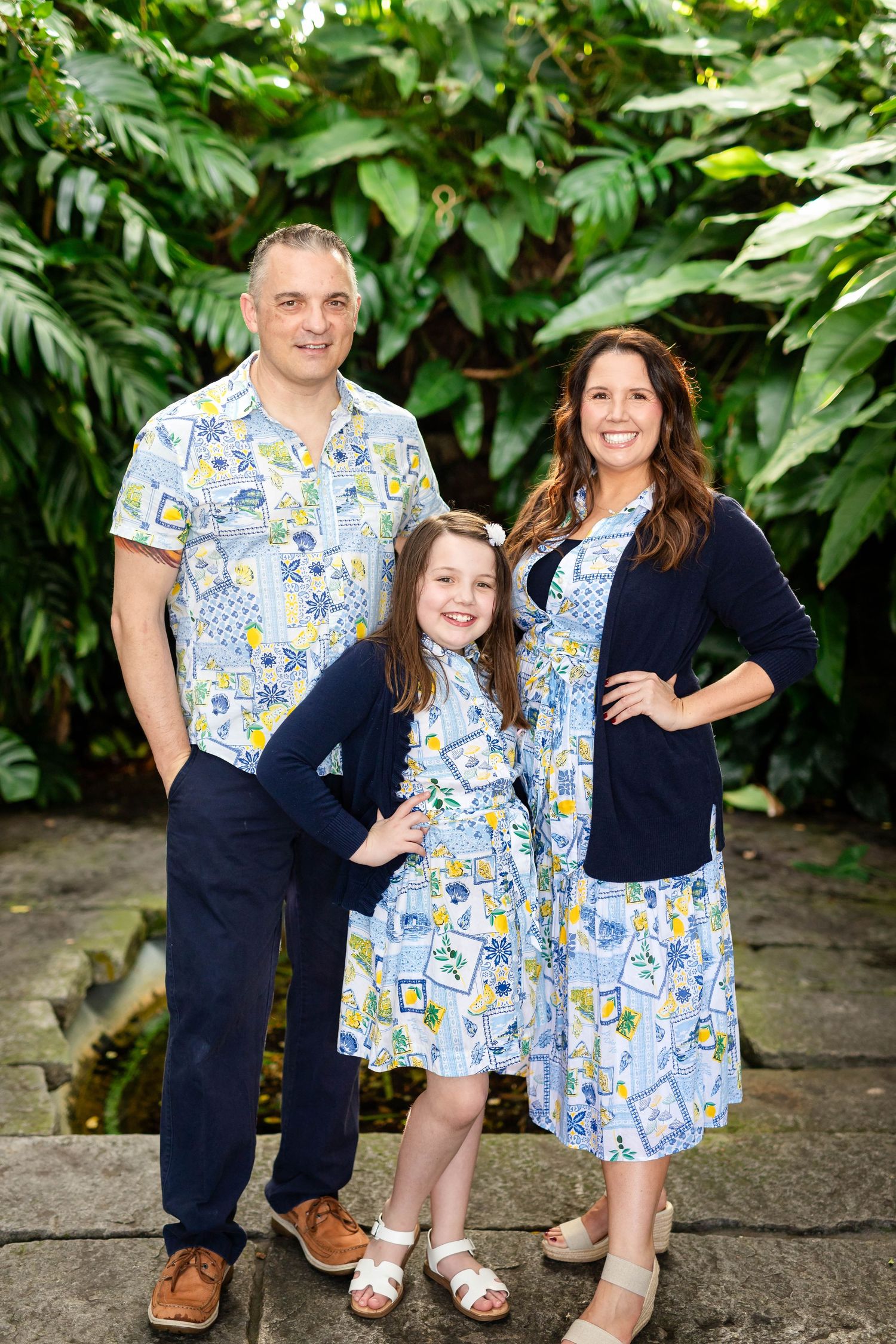 Family of three in matching blue patterned outfits poses before lush tropical green foliage backdrop at Hollis Gardens.
