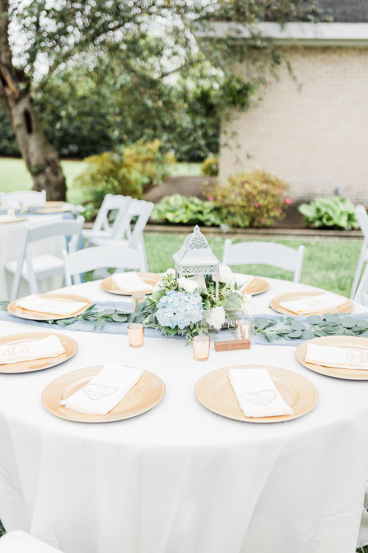Elegant outdoor wedding reception table with gold chargers, blue hydrangeas, and lantern centerpiece.