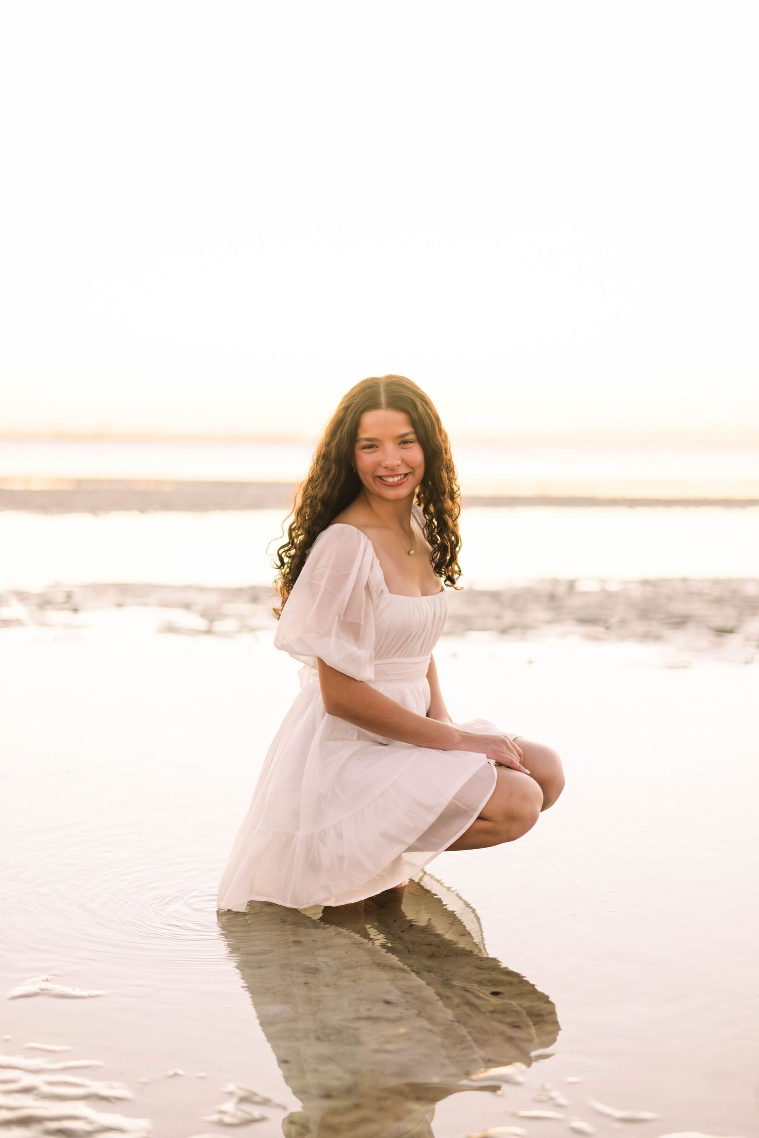 Young woman in white dress crouching on rocks at beach during golden hour sunset, smiling at camera.