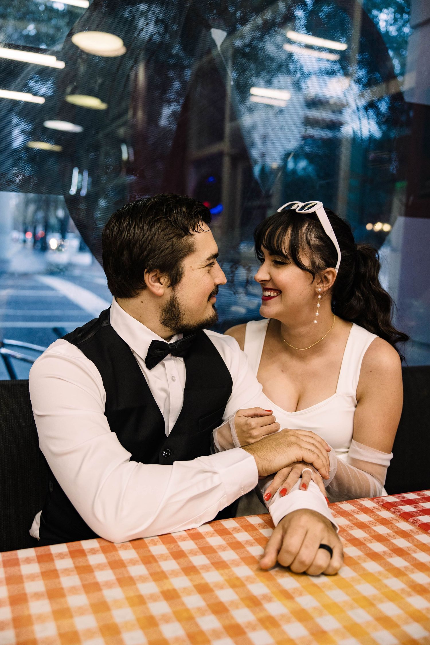 Bride and groom in retro attire laugh together at checkered tablecloth restaurant booth.