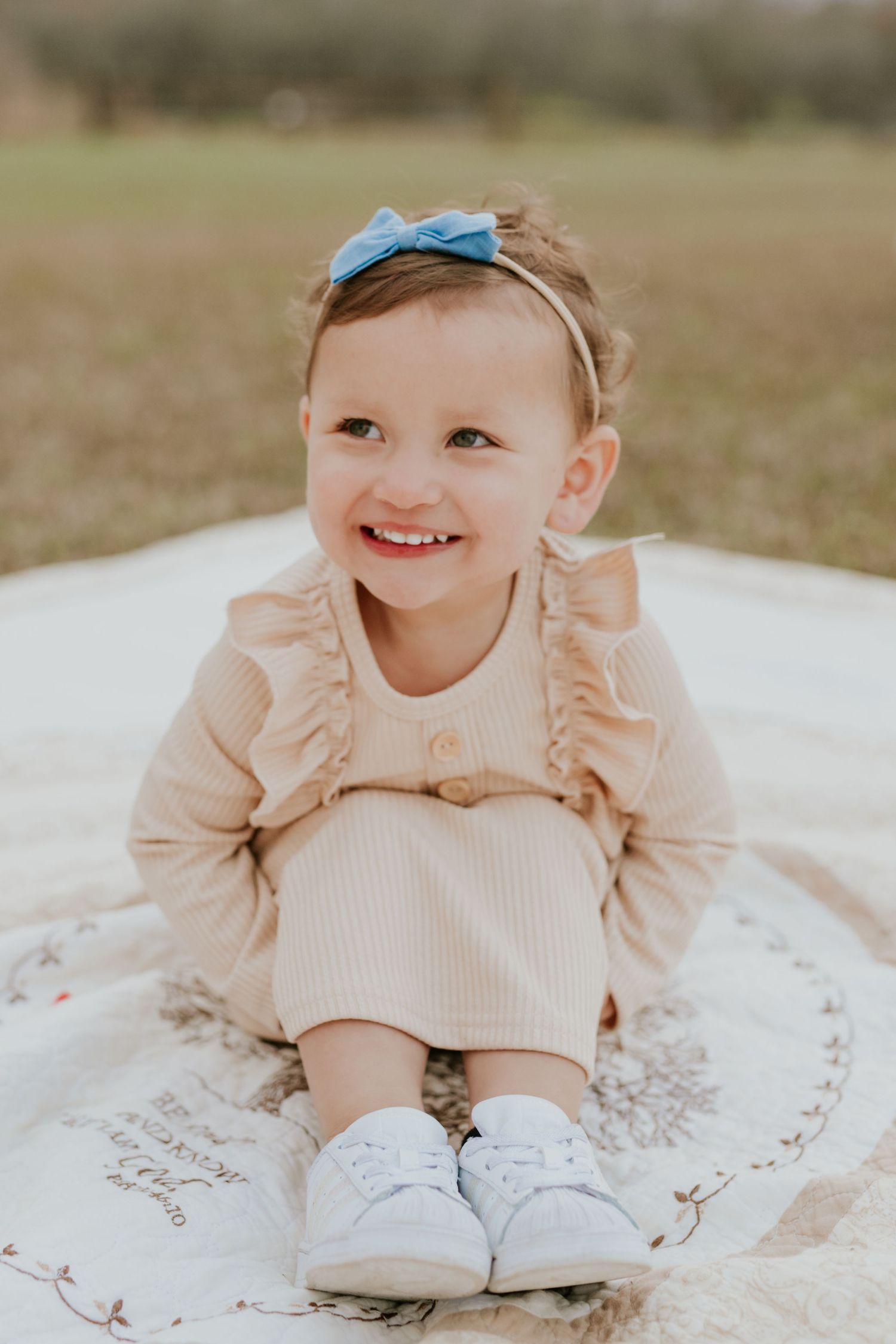 Adorable toddler girl in beige ruffle outfit with blue bow headband sitting outdoors.