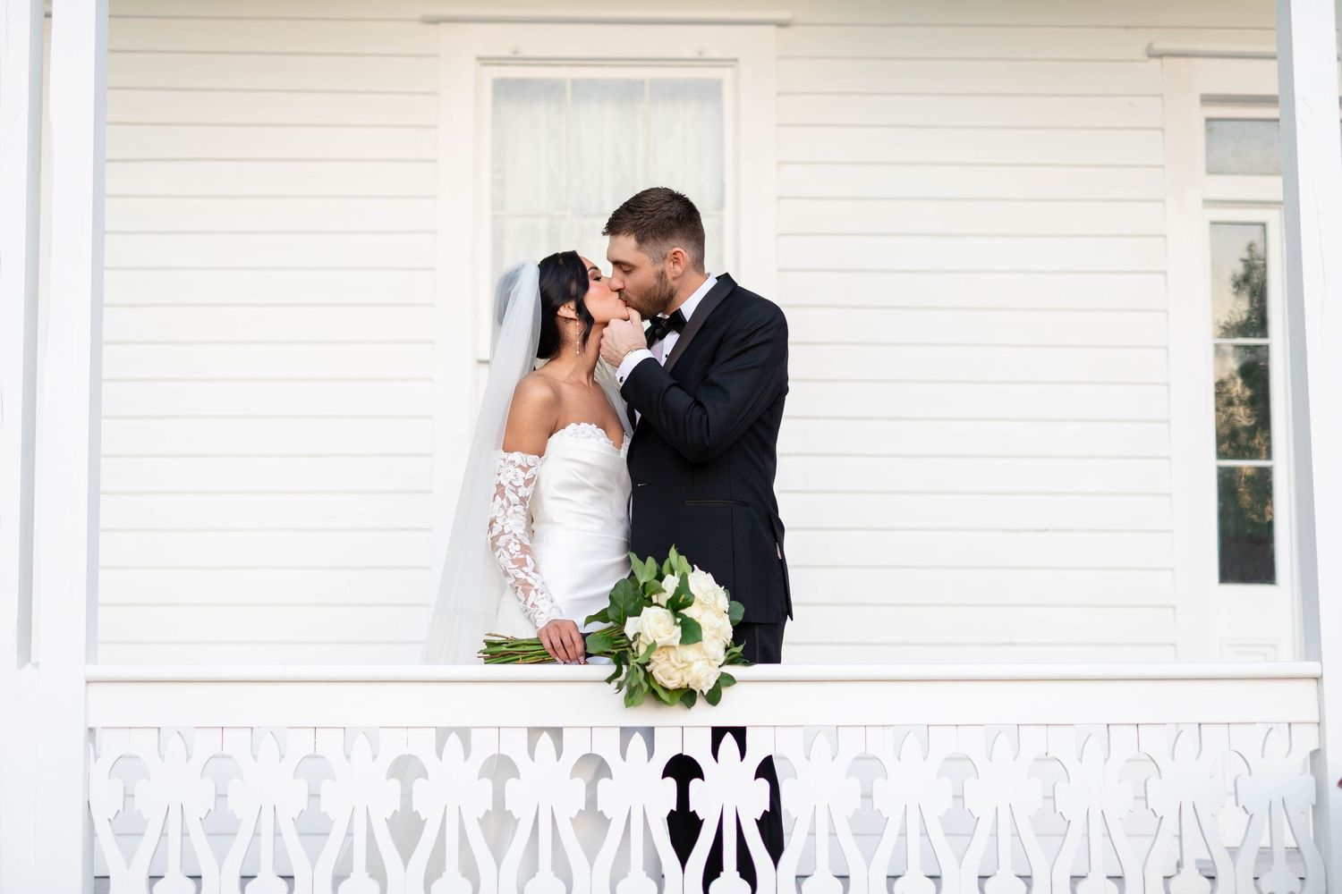 Bride and groom share a tender kiss on a white decorative balcony, bride holding a white bouquet.