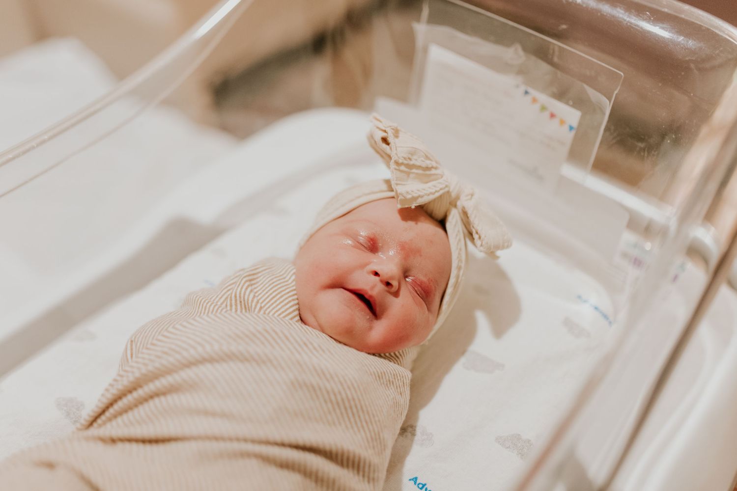 Peaceful newborn baby in beige swaddle and bow hat lying in hospital bassinet after birth.