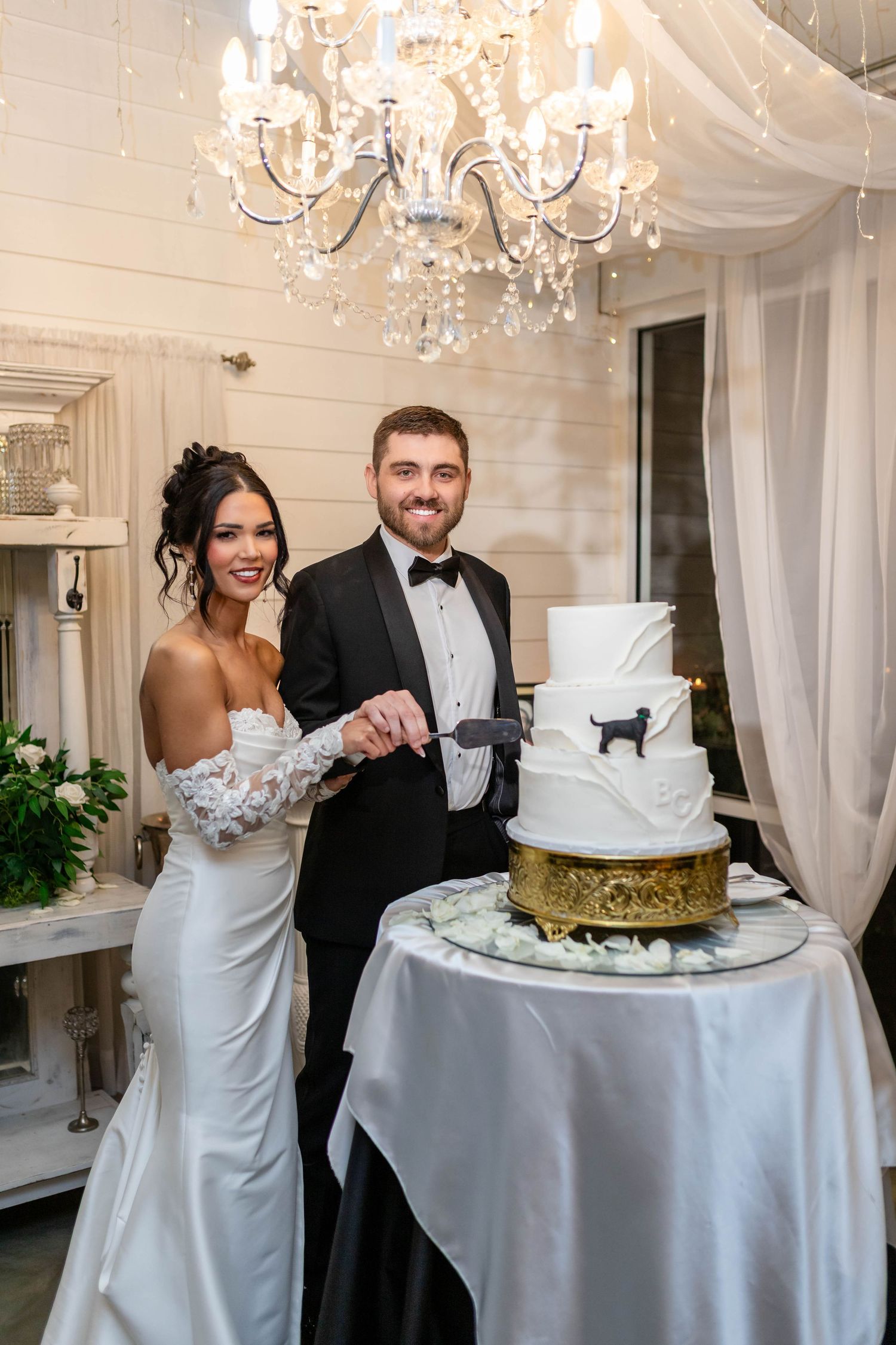 Smiling bride and groom cut their elegant three-tier white wedding cake under a crystal chandelier in Tampa.