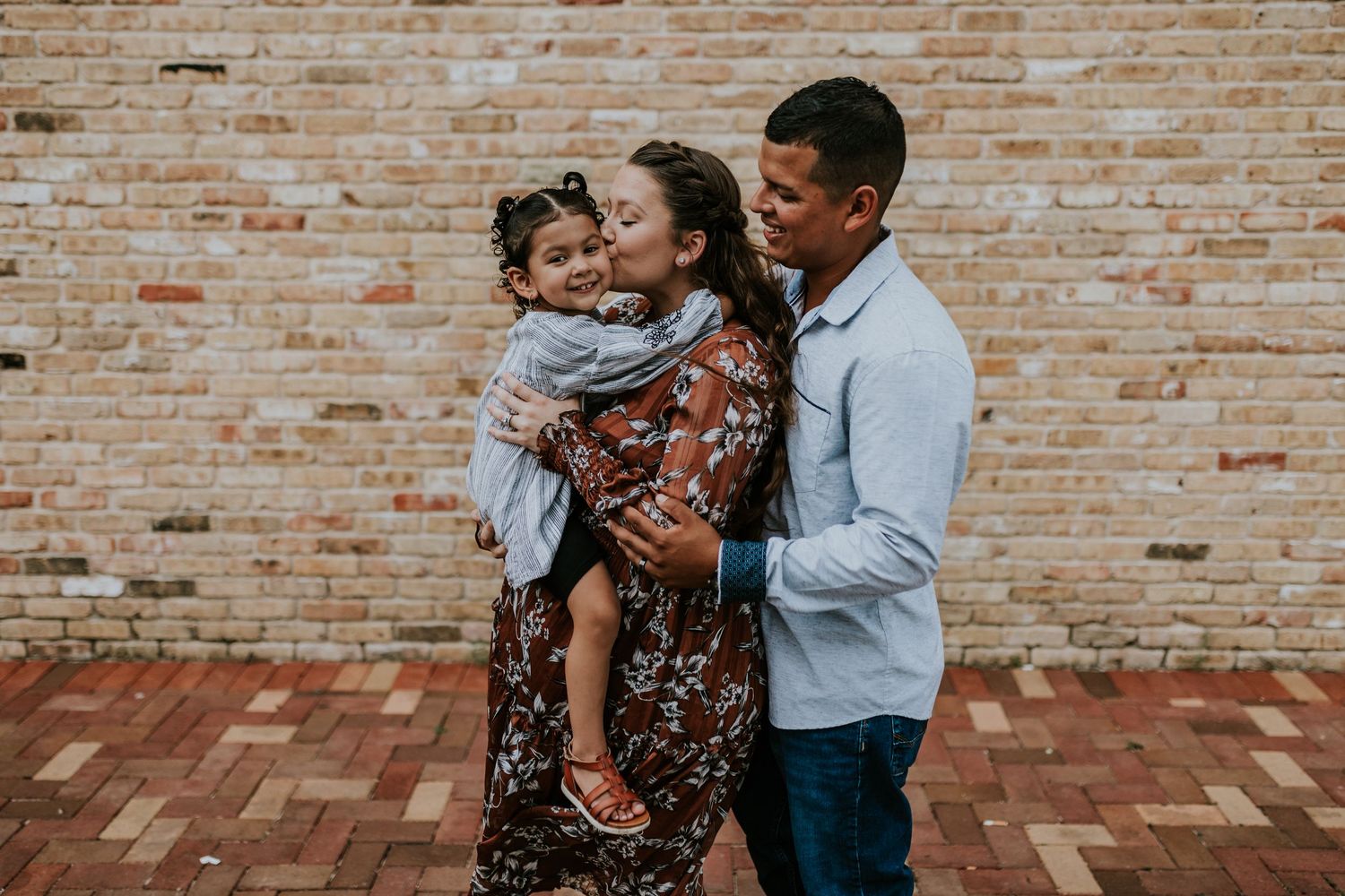 A joyful family shares a warm embrace against a rustic brick wall, with parents kissing their young child.