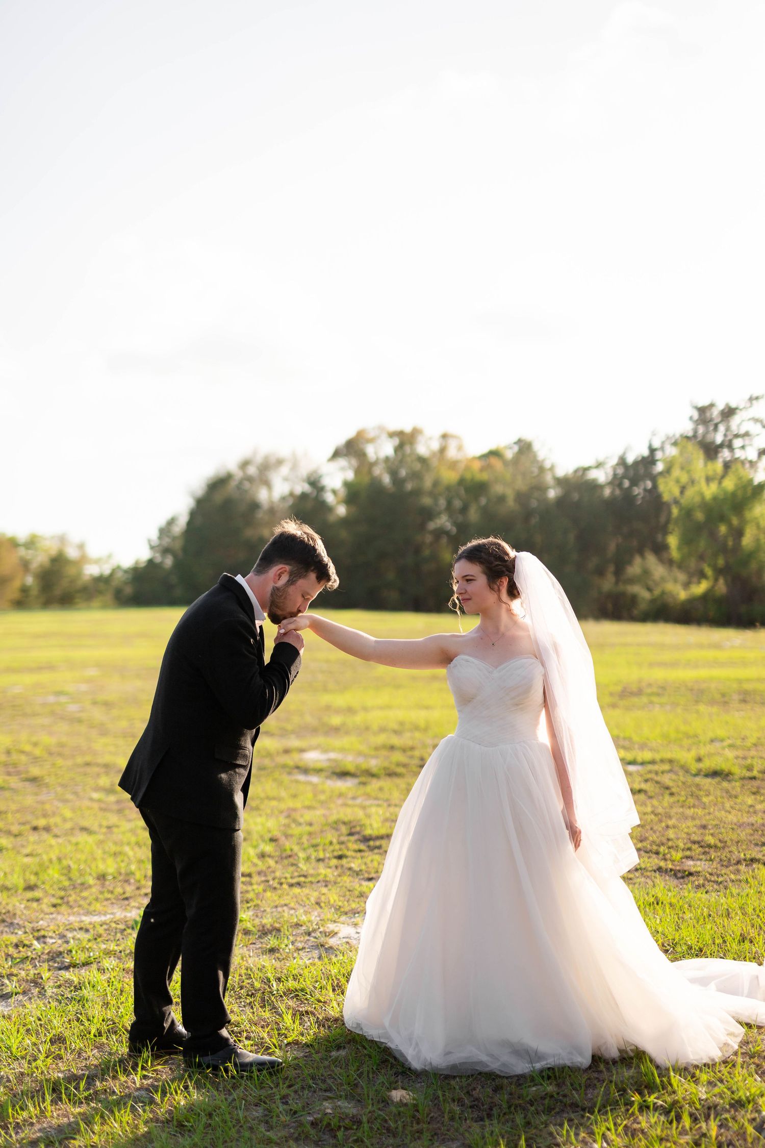 Groom tenderly kissing bride's hand while she smiles in golden hour outdoor wedding photo.