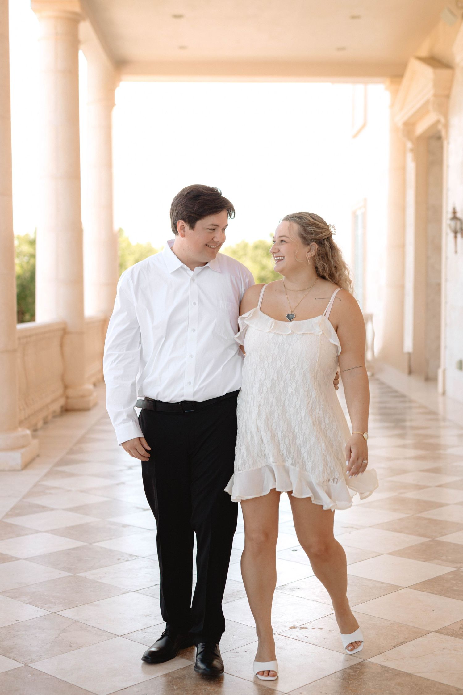 Young couple walking and smiling at each other in elegant attire along a sunlit columned corridor.