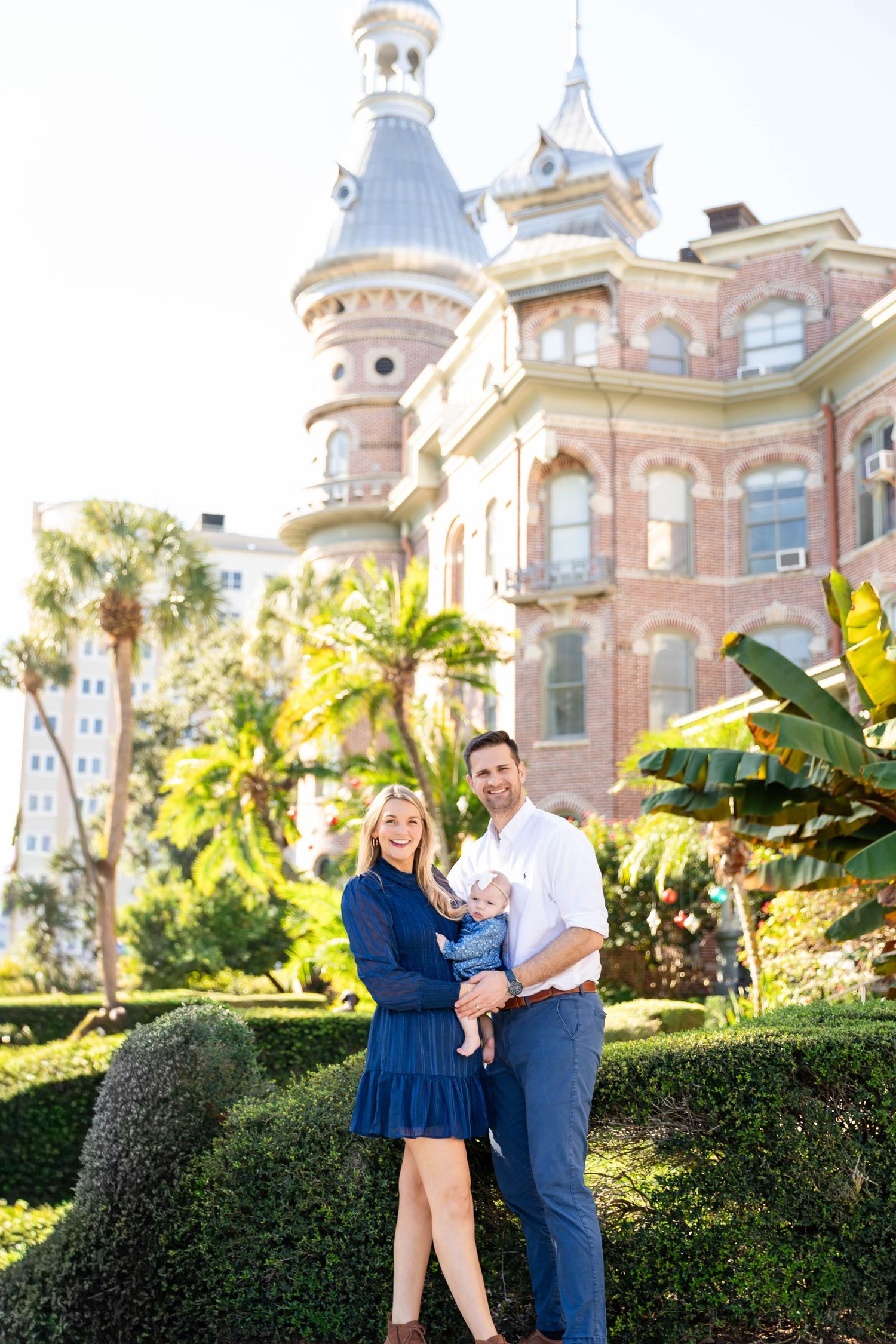Joyful family of three poses with laughing baby before ornate Victorian brick tower building at the University of Tampa..