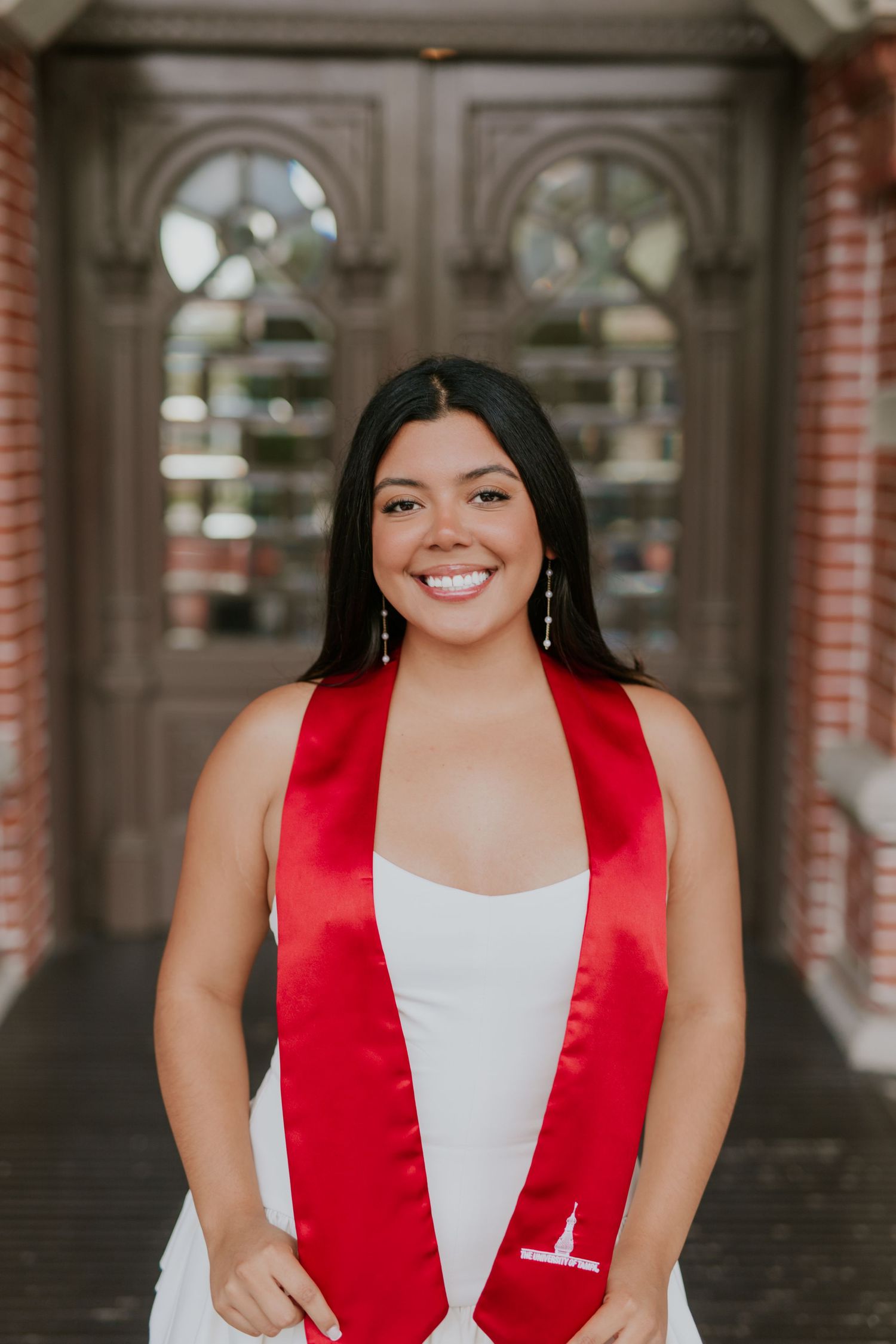 Smiling graduate in white dress and red satin stole poses in front of ornate wooden doors.