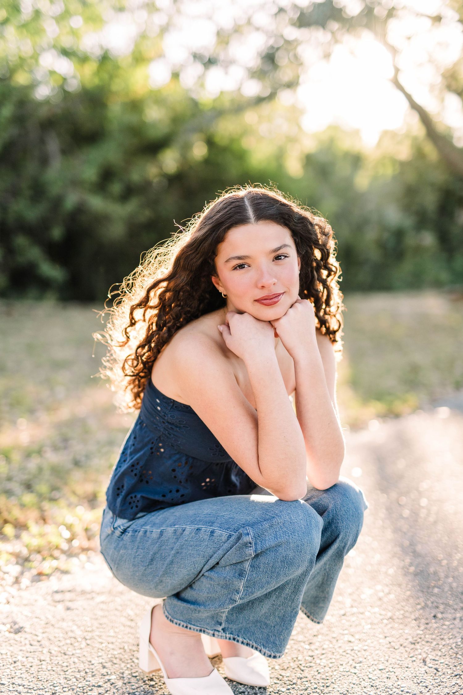 Young woman with curly hair crouching on a path, wearing jeans and a navy top, smiling in golden hour light.
