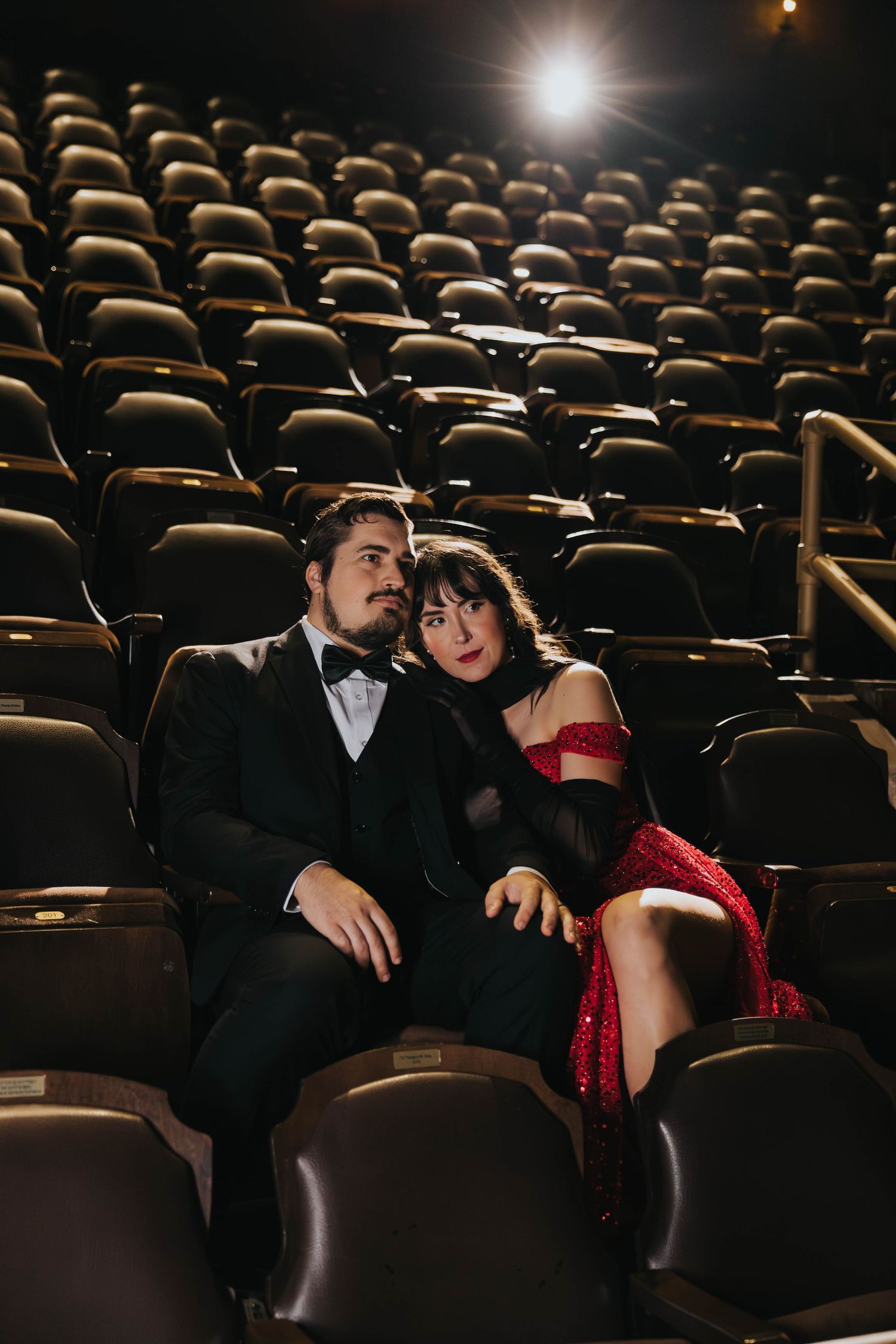 Elegant couple in formal wear sitting together in empty Tampa theater seats in dim light.