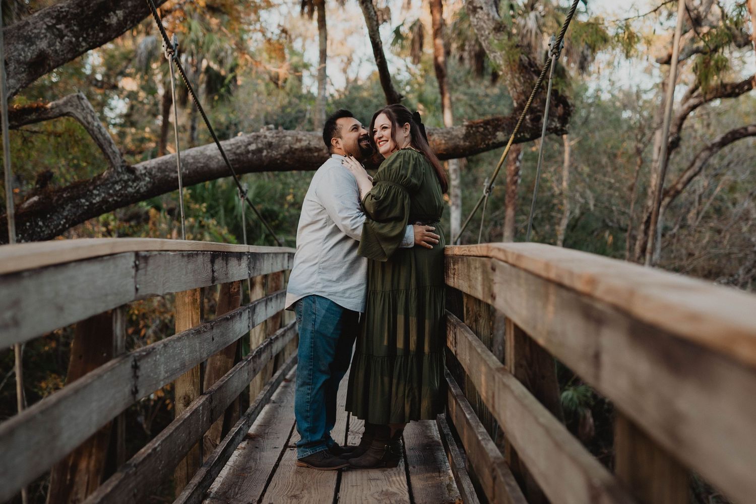 Couple embracing on a wooden bridge surrounded by lush trees in a romantic outdoor setting.