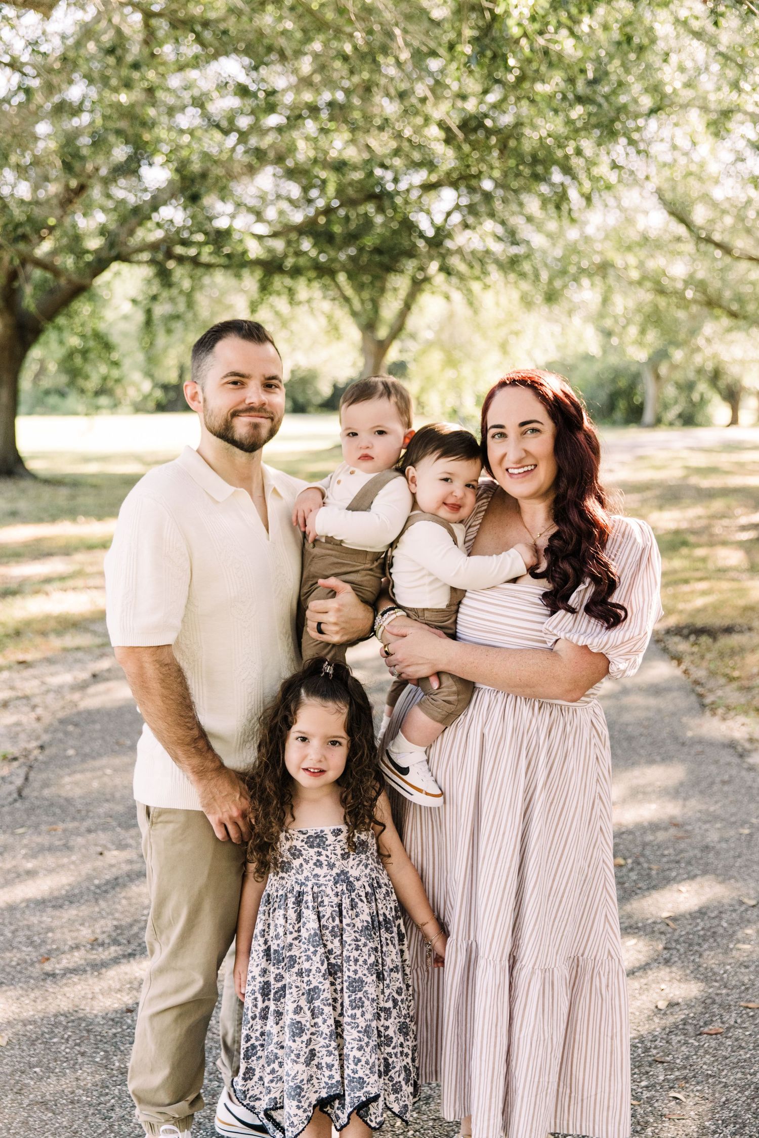 Family of five with twin babies and curly-haired girl posing outdoors under trees at Cypress Pointe Park.