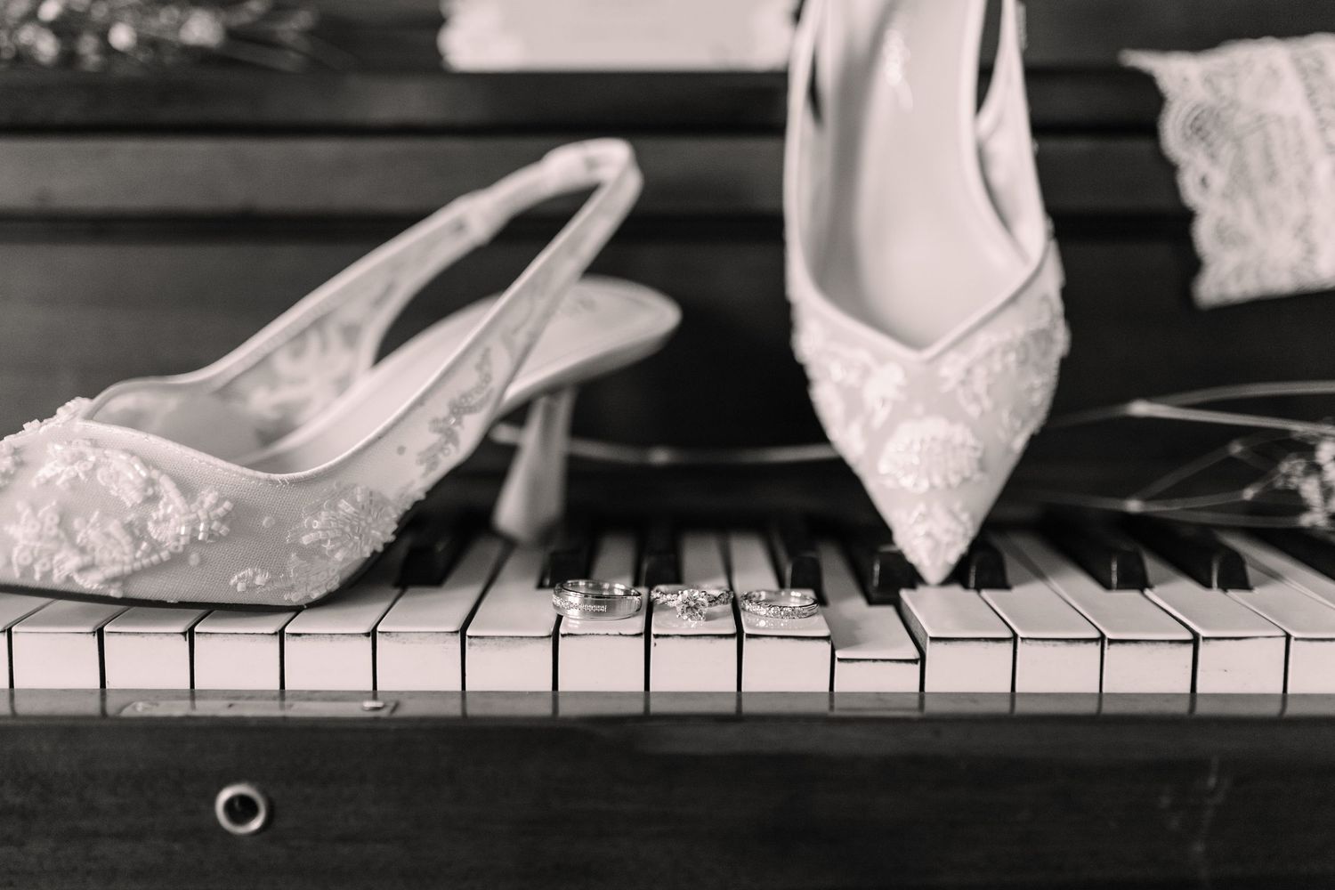 Black and white photo of elegant lace bridal heels resting on piano keys with wedding rings nearby.