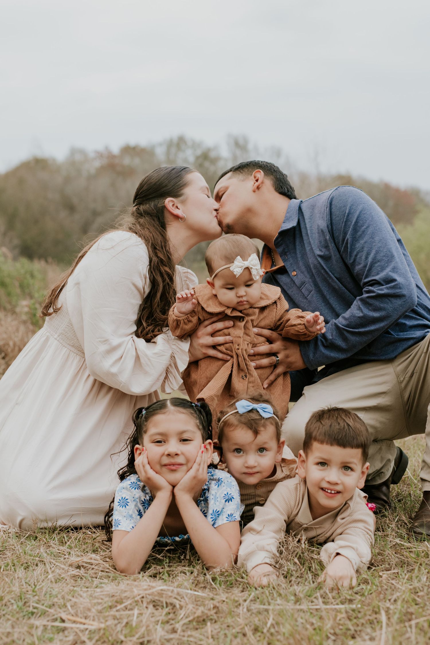 Family of six posing outdoors, parents kissing over baby while three children smile in front.