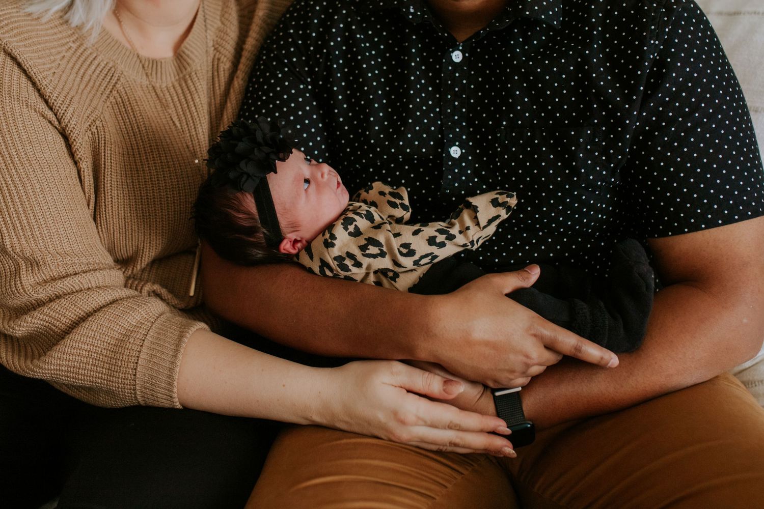 Interracial couple cradles their newborn dressed in leopard print in a tender close-up moment.