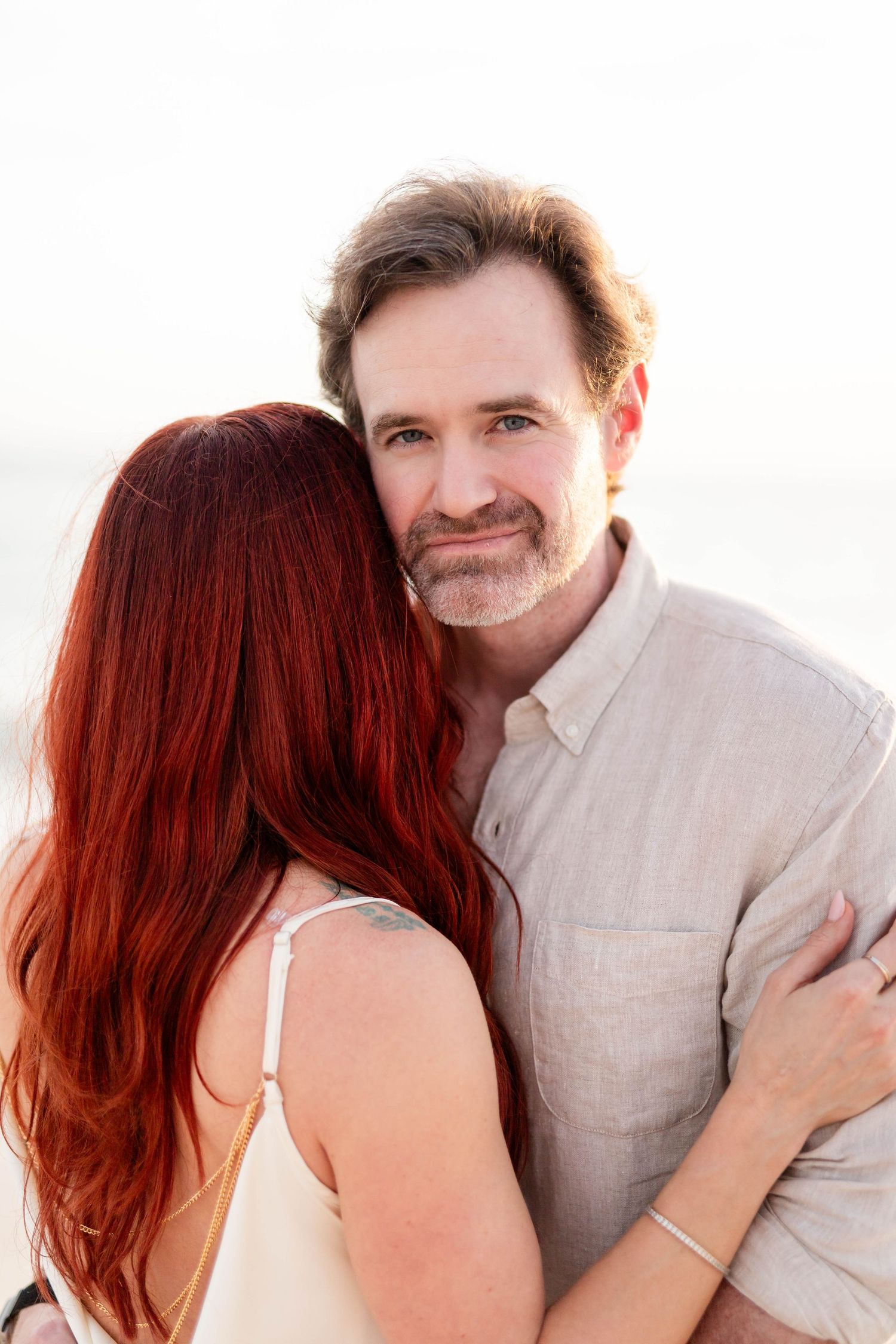Man embracing red-haired woman from behind, smiling at camera in a bright, softly lit outdoor portrait at Honeymoon Island.