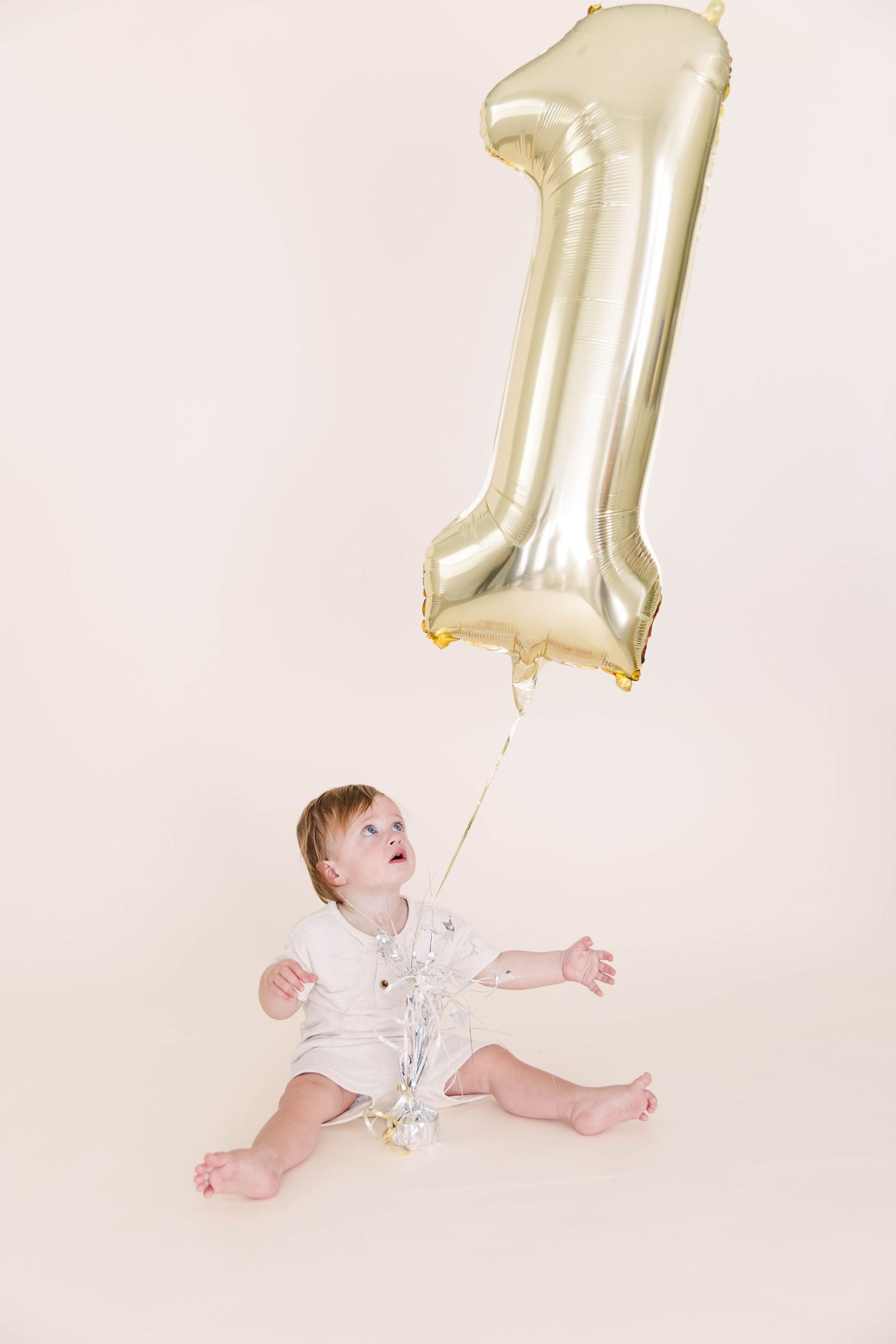A curious baby in white looks up at a gold number one birthday balloon.