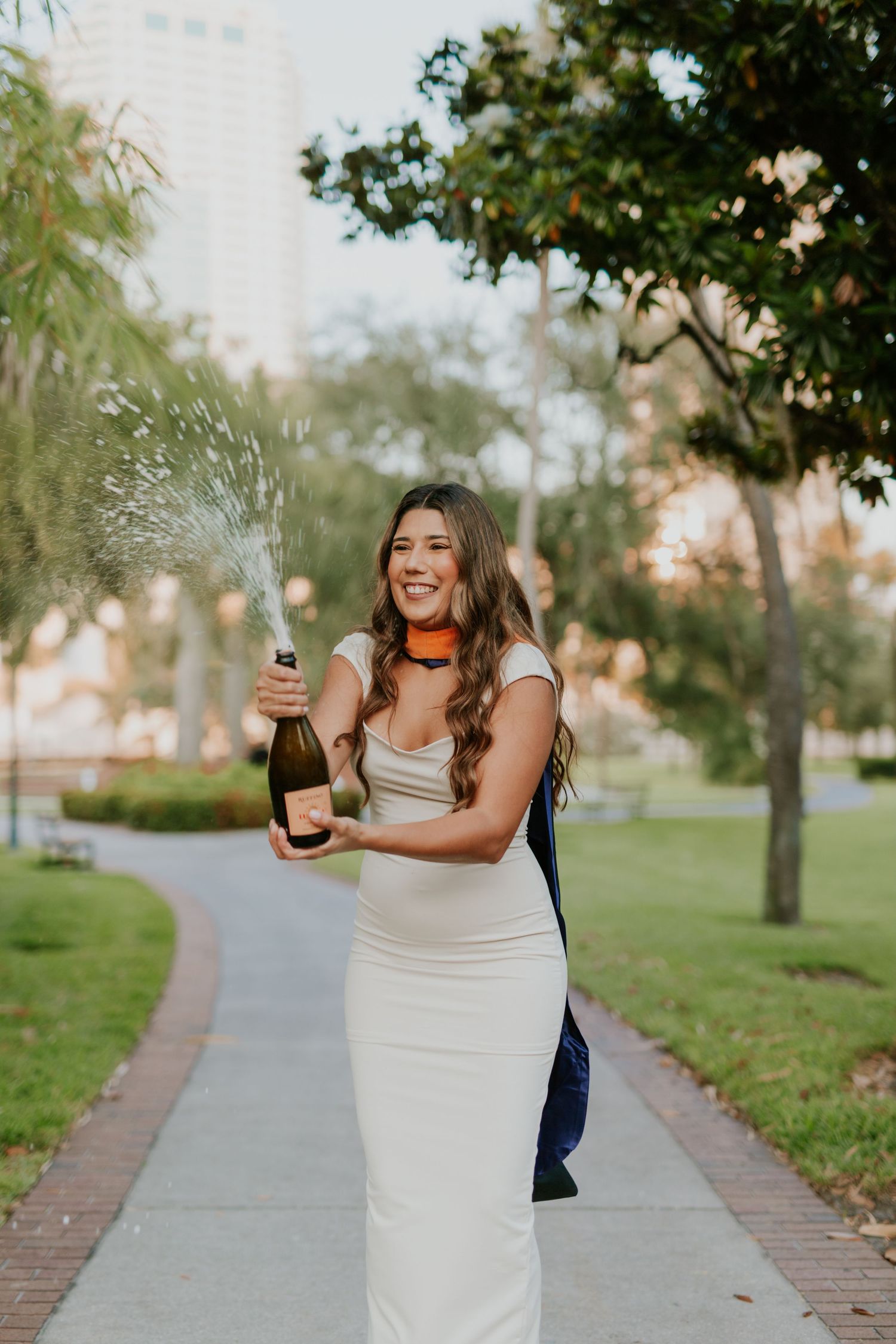 Young woman in white dress joyfully popping champagne bottle outdoors in a lush green park setting.