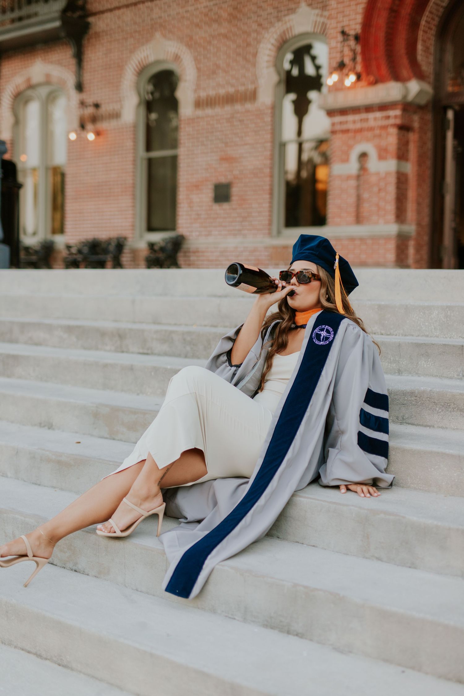 Woman in graduation cap and gown sitting on steps, drinking from bottle, celebrating academic achievement.
