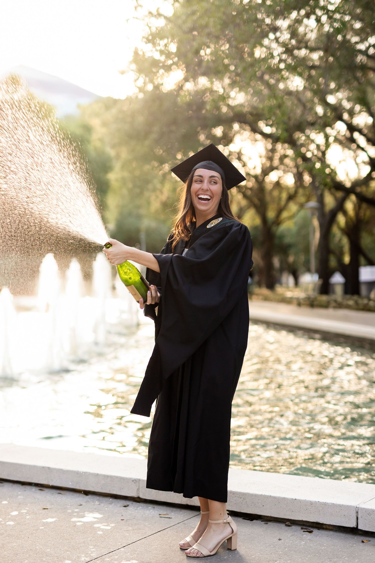 Joyful graduate in black cap and gown spraying champagne bottle outdoors near a fountain at sunset at the University of South Florida.
