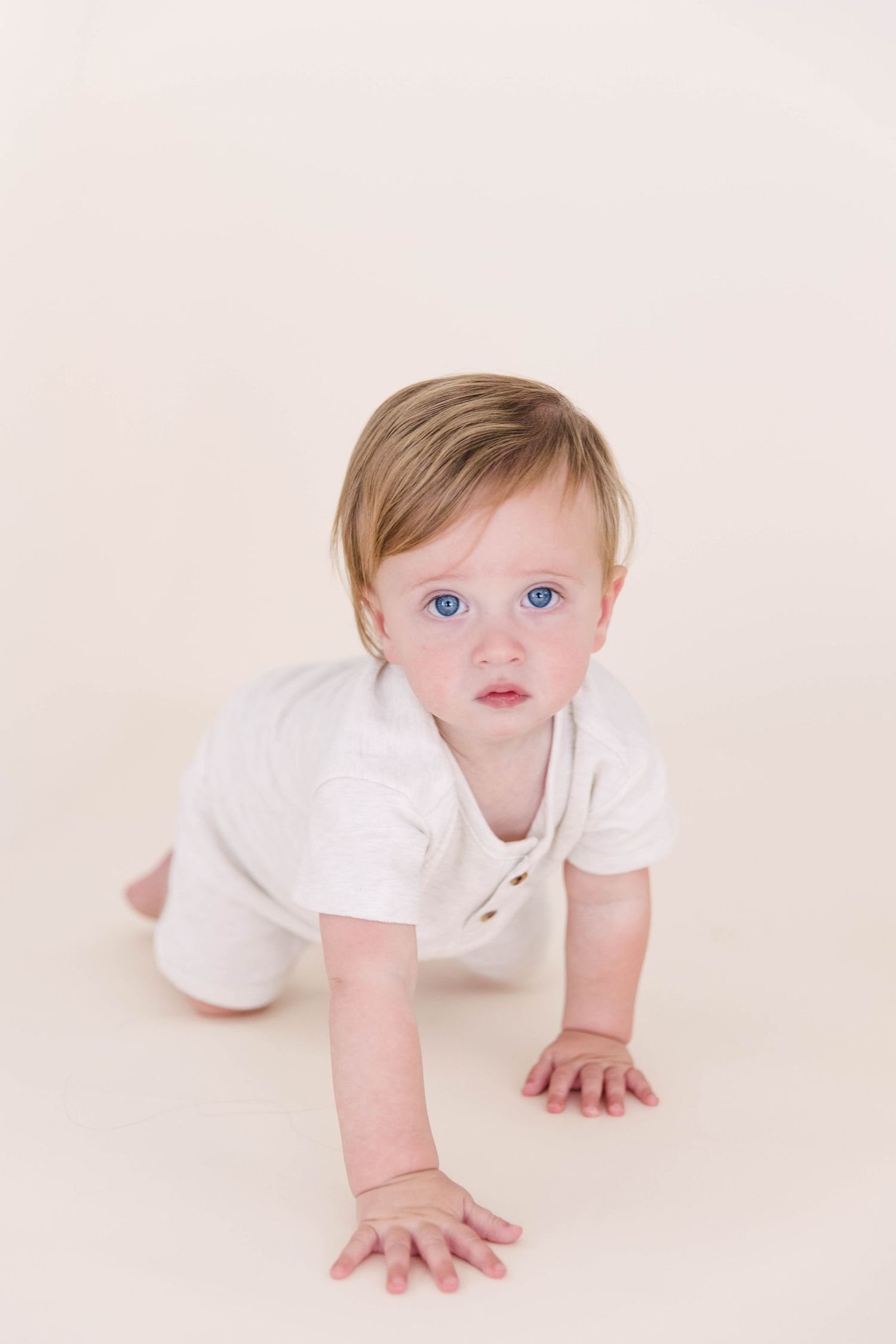 Blue-eyed baby in white onesie crawling on cream floor.