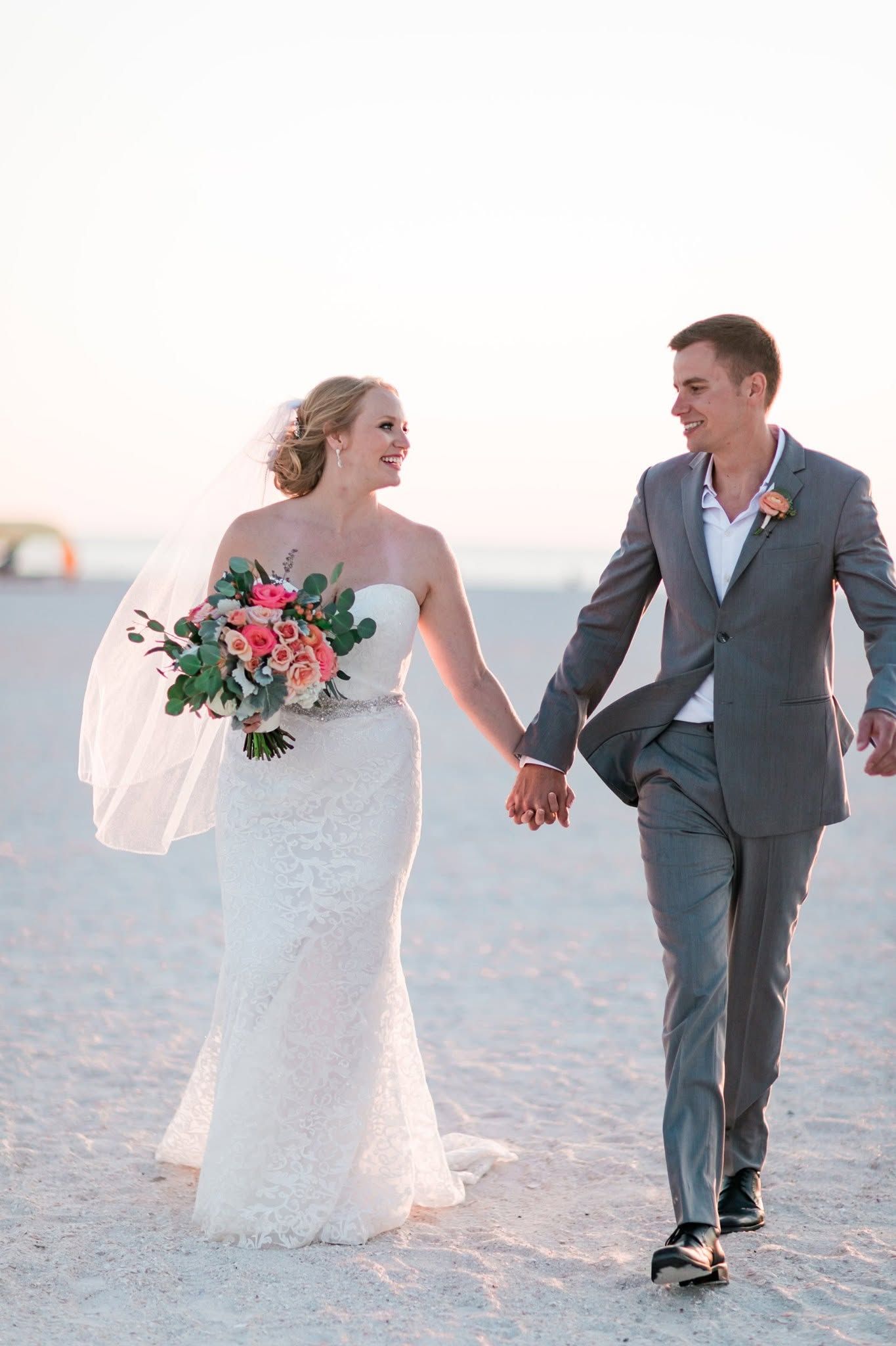 Bride and groom walk hand in hand on sandy beach at golden sunset holding floral bouquet.