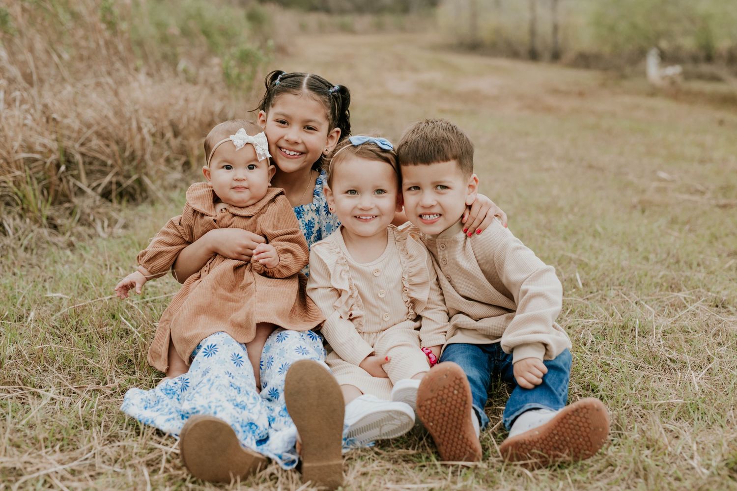 Four young siblings sitting together smiling in grassy outdoor field.