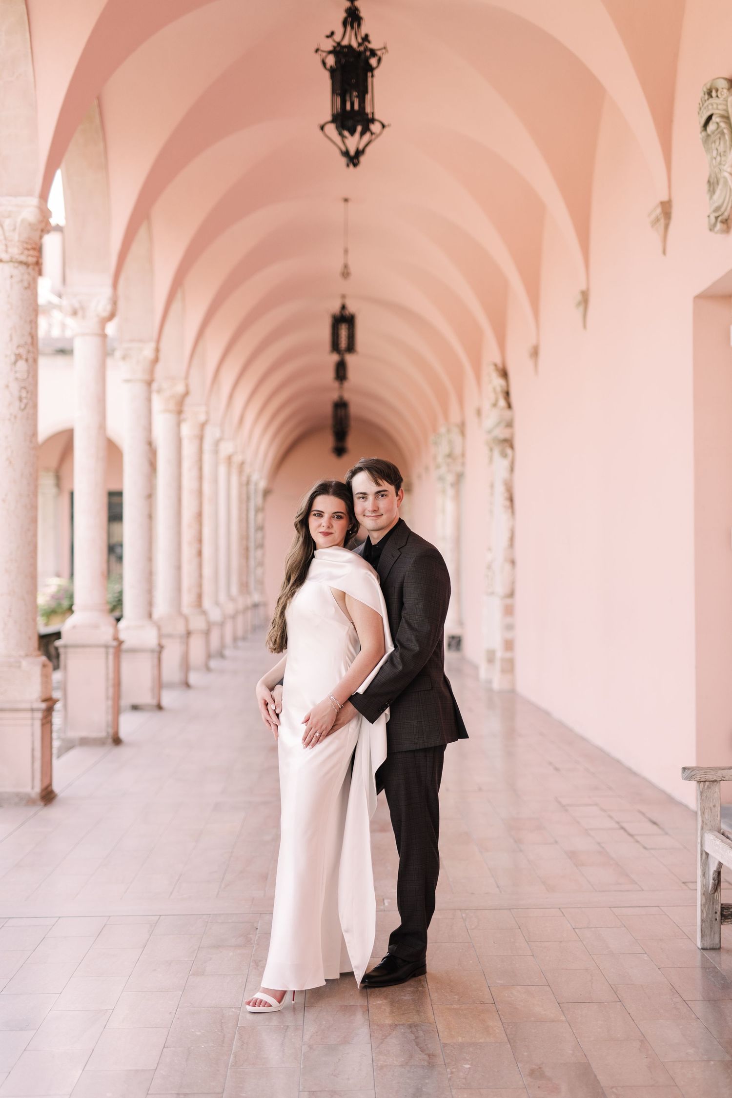 Elegant bride in white gown and groom in black suit pose in pink arched colonnade on wedding day at The Ringling Museum.