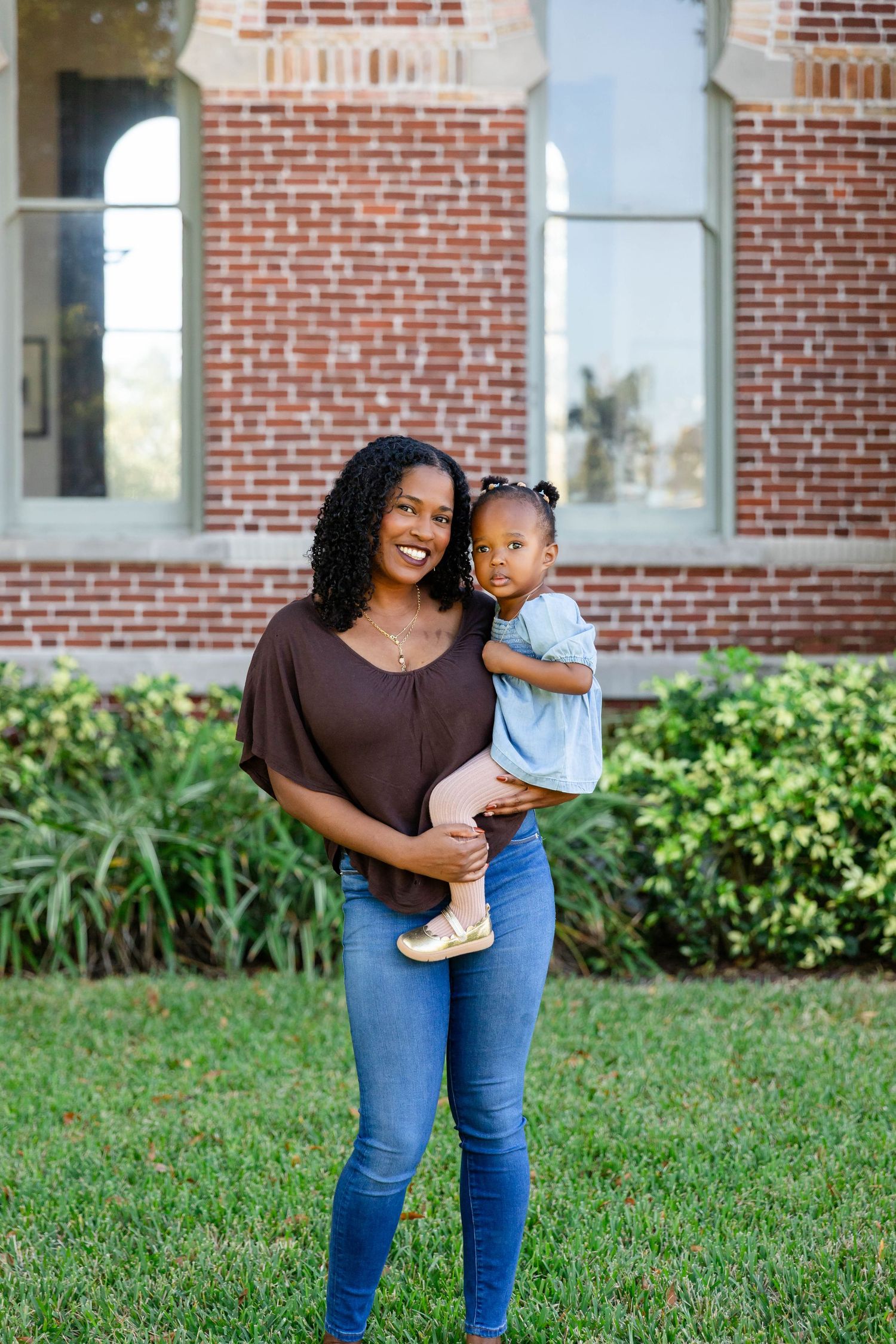 Smiling Black mother holds young toddler daughter while standing on green lawn near brick building at the University of Tampa.
