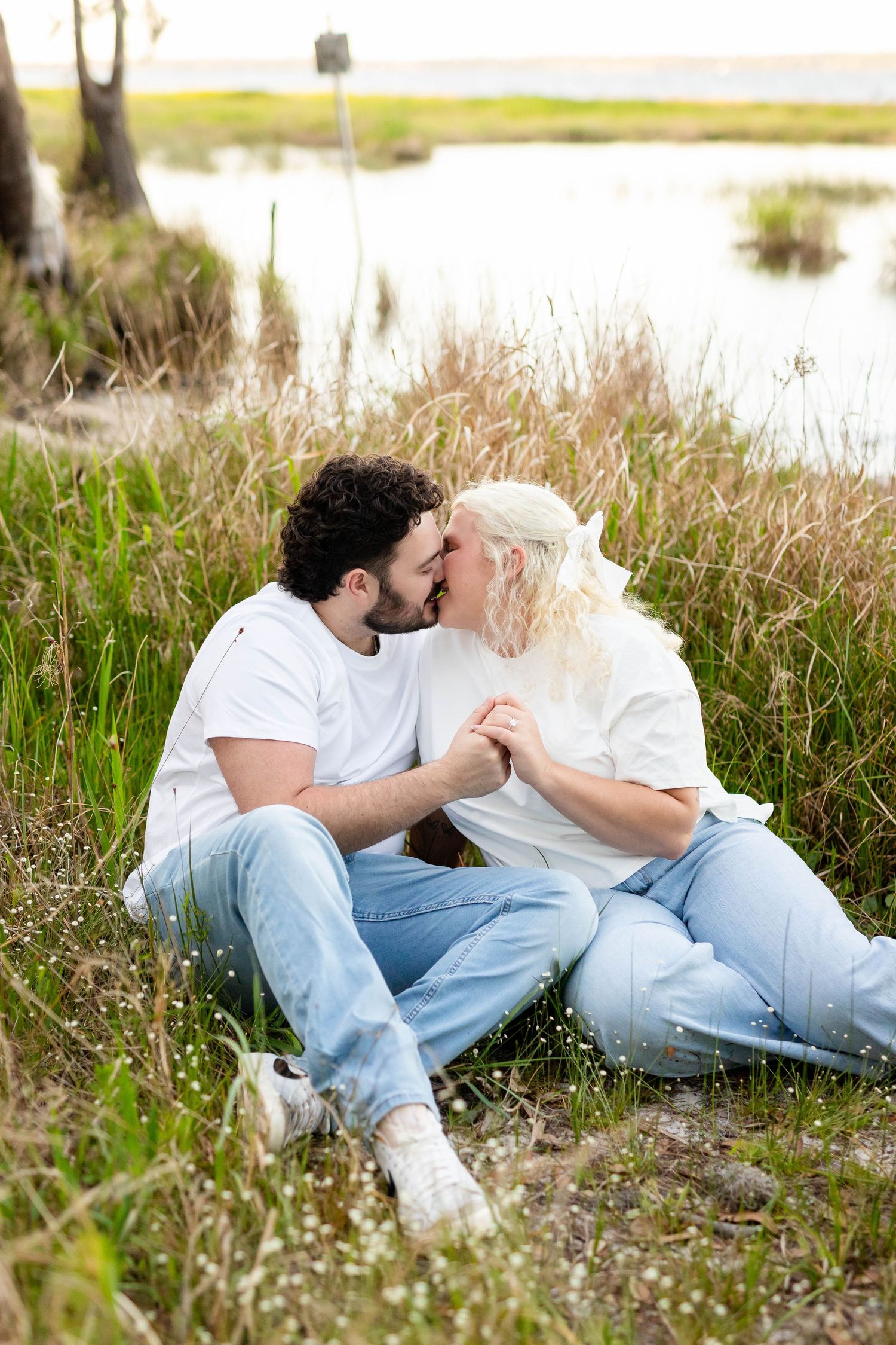 Engaged couple kissing while sitting in tall grass beside a serene lake, both wearing casual white outfits.
