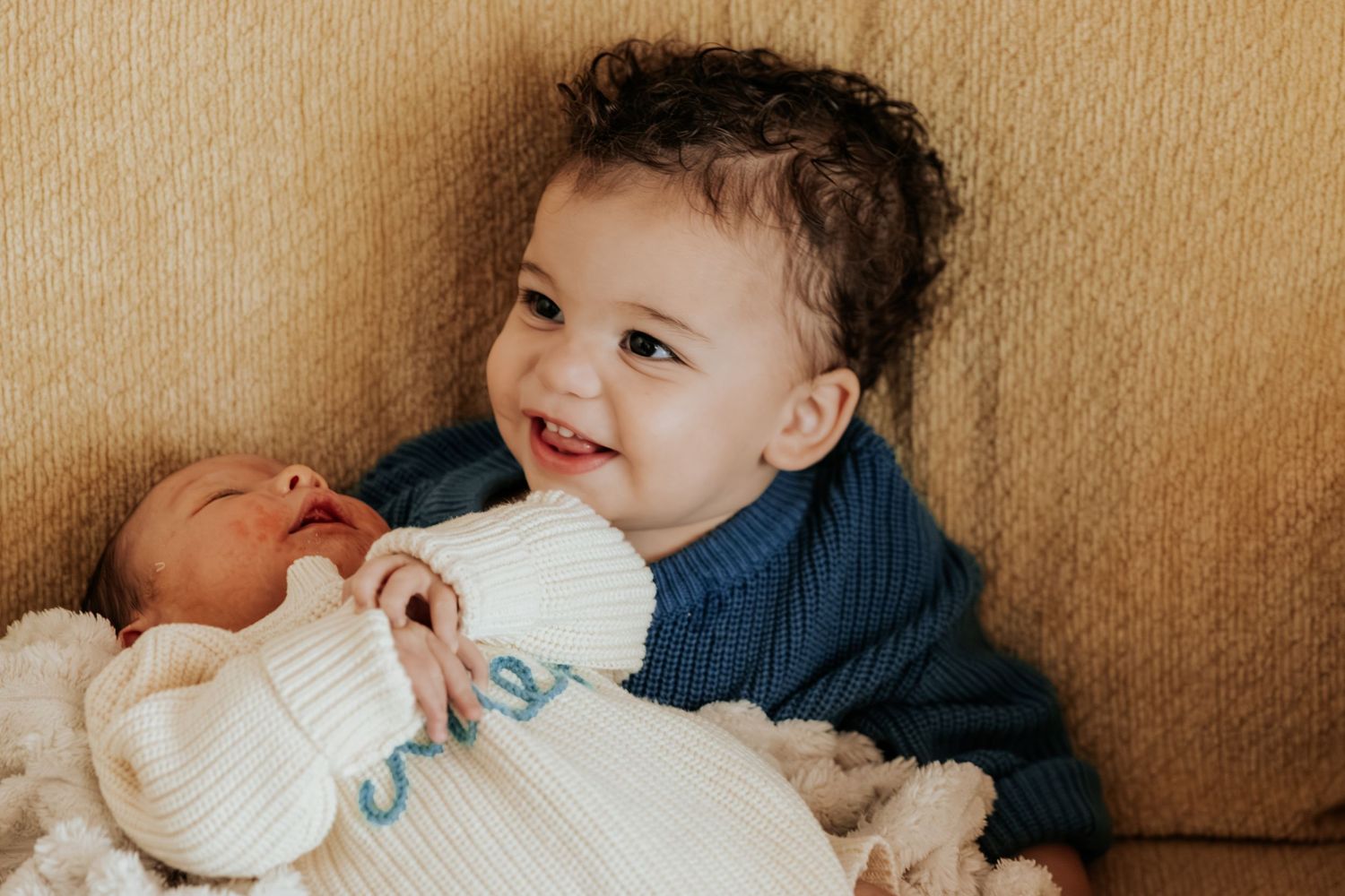 Smiling toddler in blue sweater proudly holds swaddled newborn sibling on a couch.