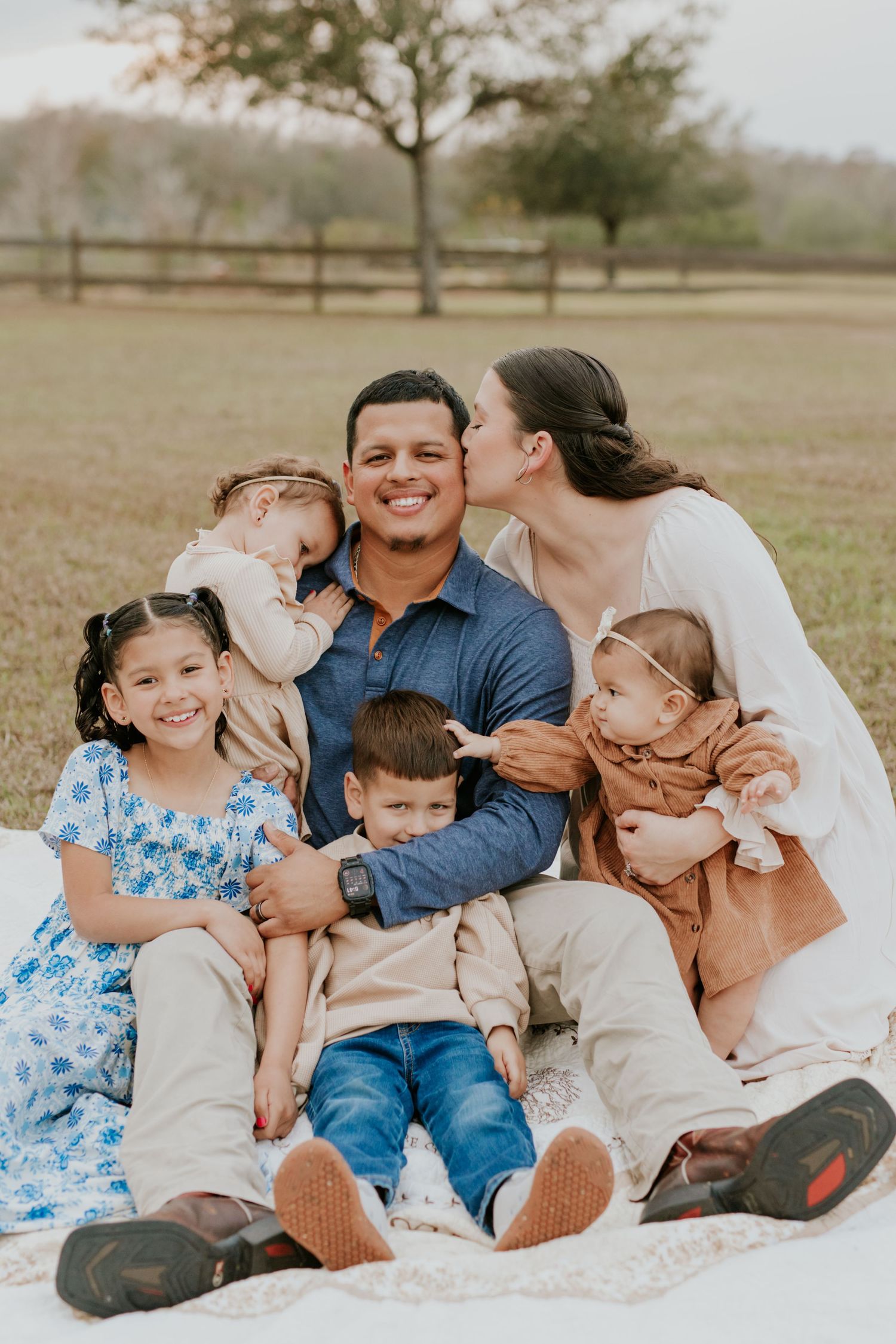 Happy family of six sitting on blanket outdoors, mother kissing father on cheek.