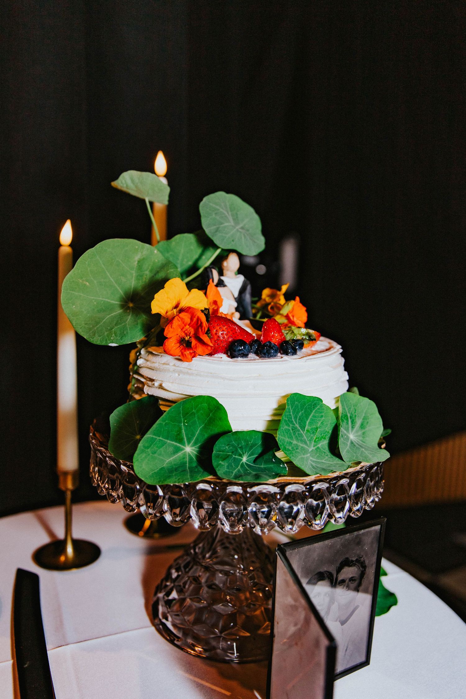 Elegant two-tier wedding cake with fresh flowers, berries, and green leaves on a crystal cake stand at the Floridian Social.