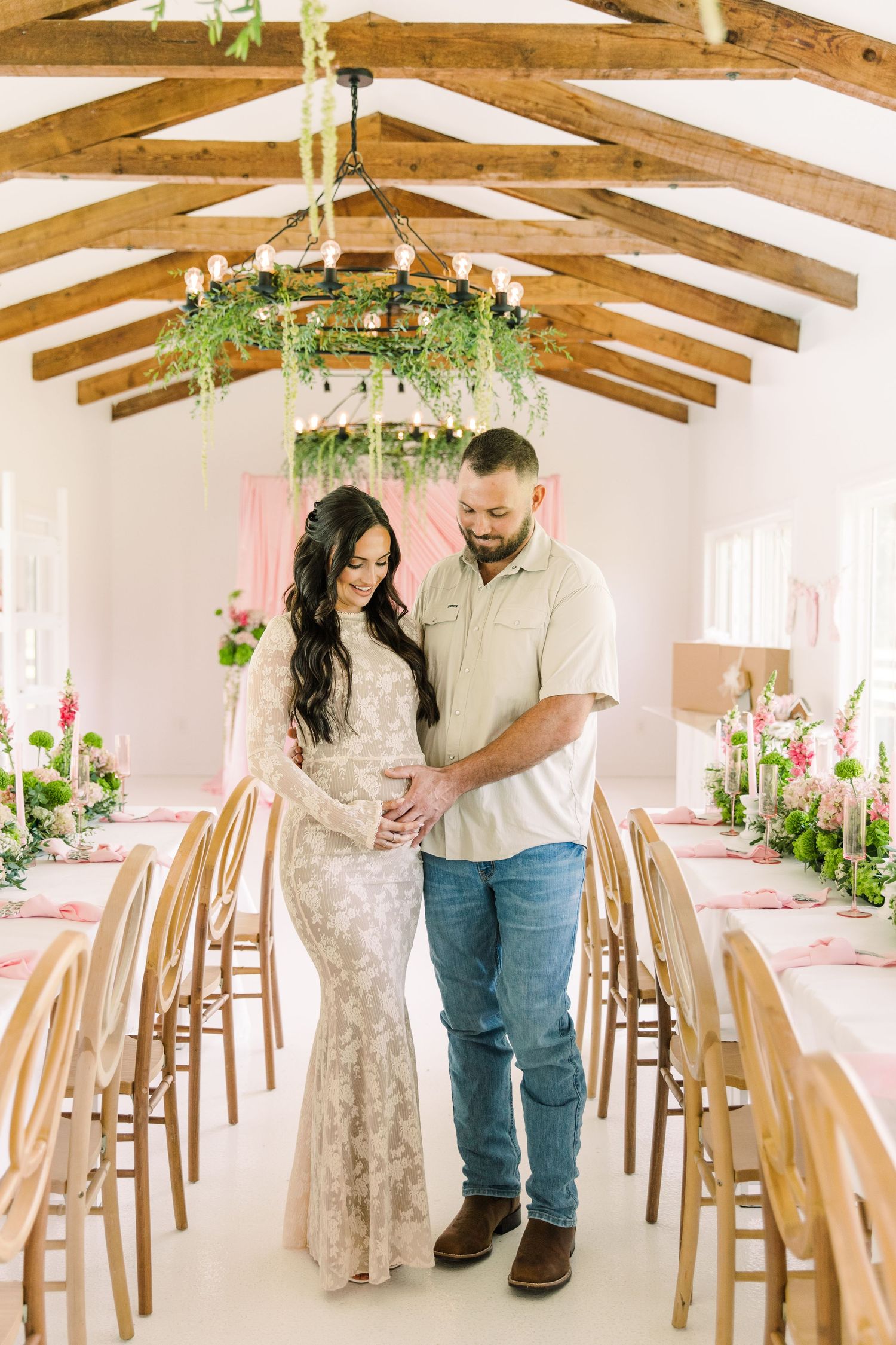 Couple tenderly holds pregnant belly in decorated event hall with wood beam ceiling and pink floral arrangements at the Colonial at Sunset..