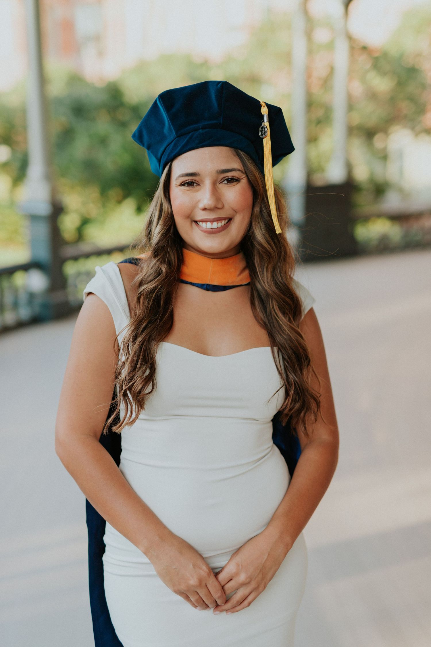 Smiling young woman in white dress and navy graduation cap with gold tassel poses outdoors for graduation photos.