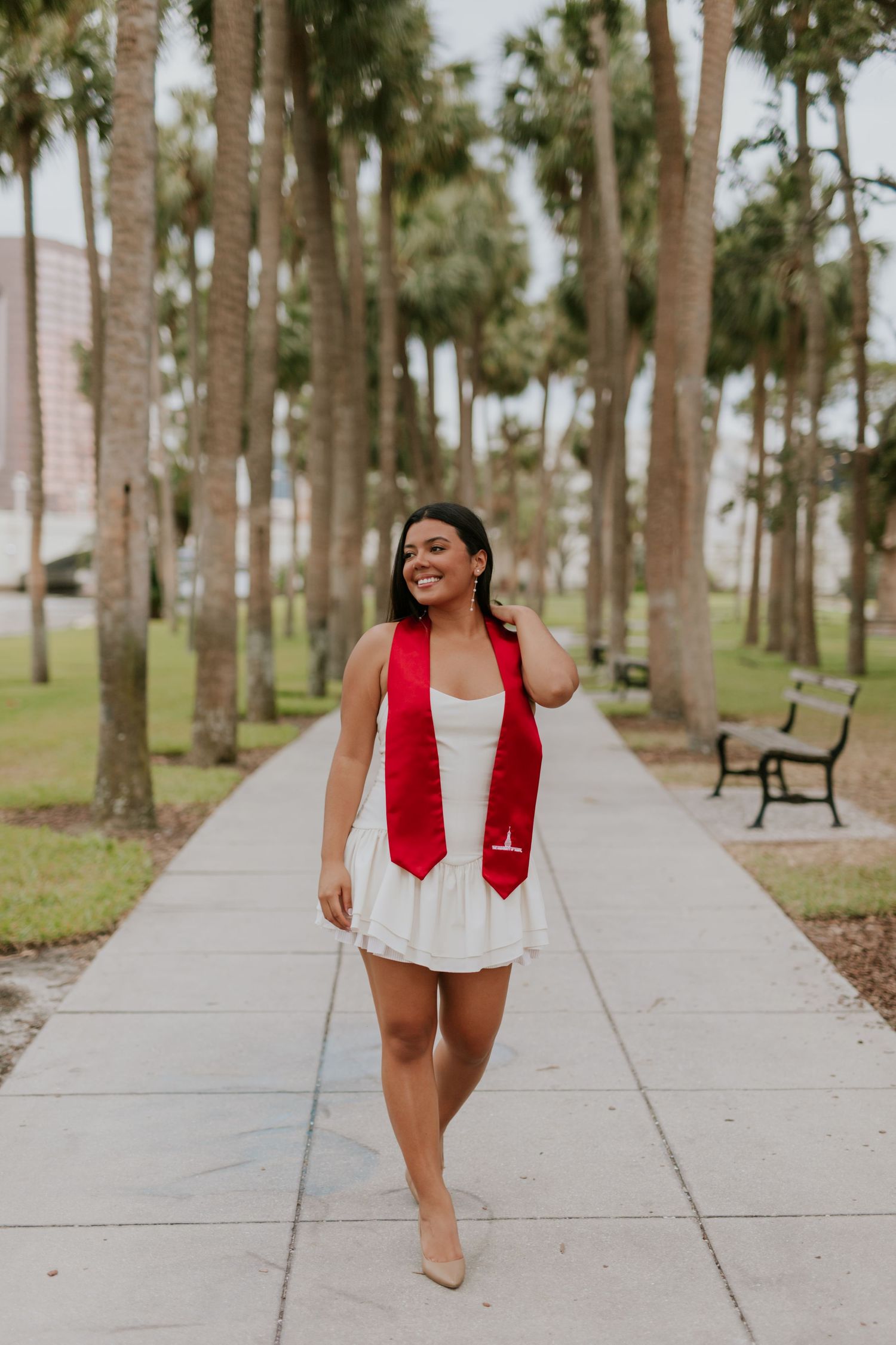Young woman in white dress and red graduation stole walks confidently along palm tree-lined pathway.