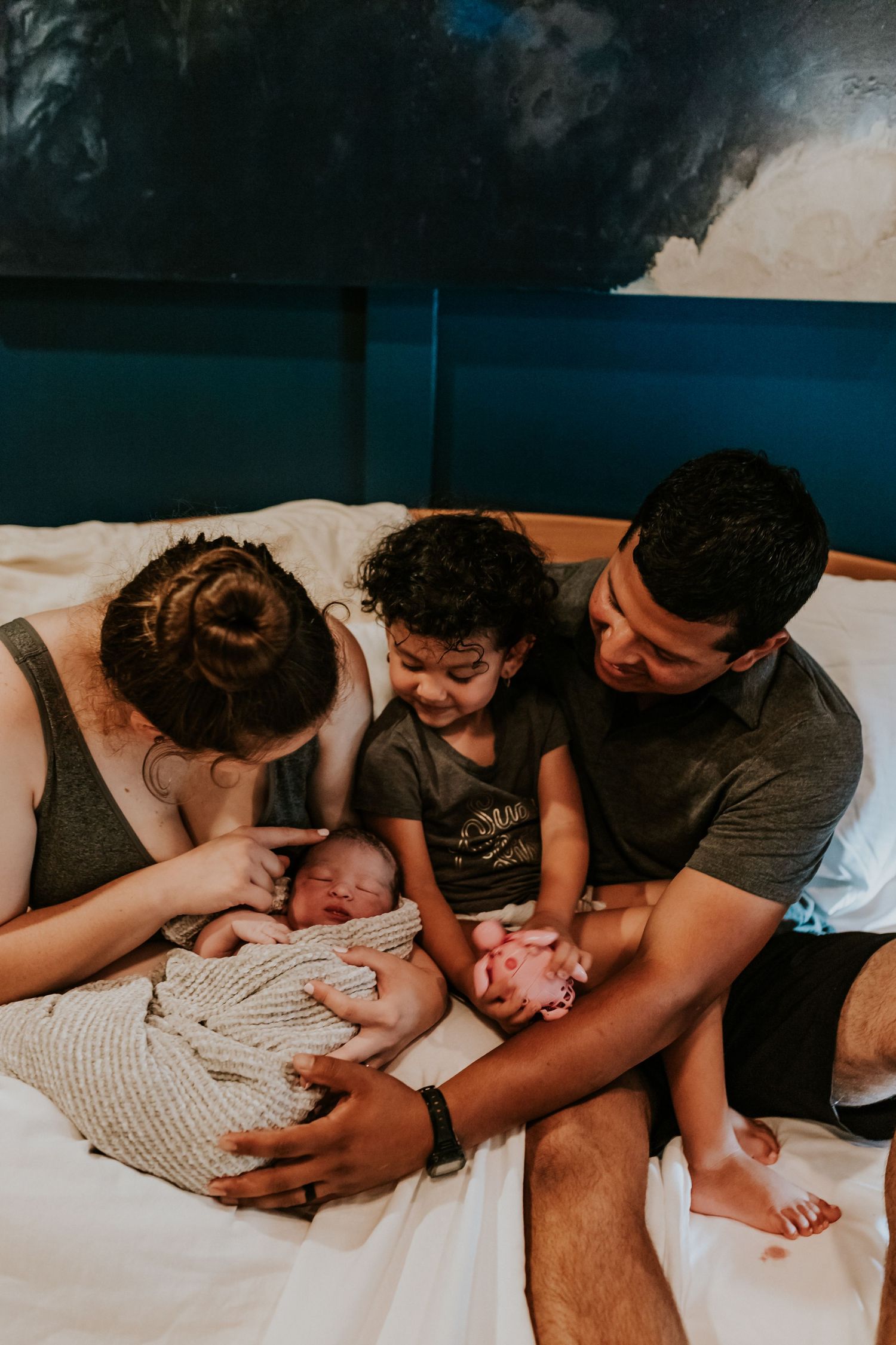 A family of four gathers on a hospital bed to welcome and admire their newborn baby together.