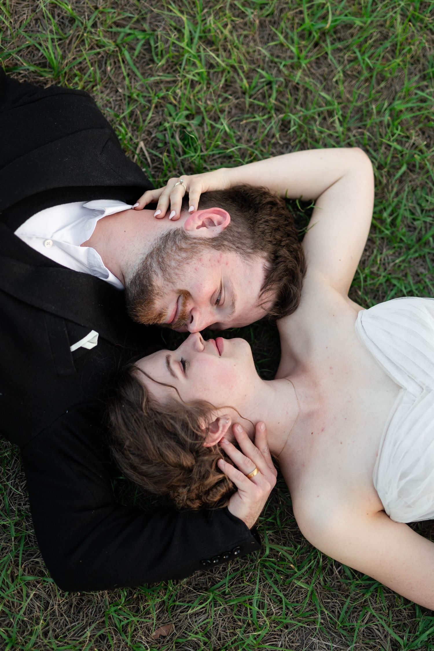 Bride and groom lying in grass sharing a romantic kiss during outdoor wedding portraits.