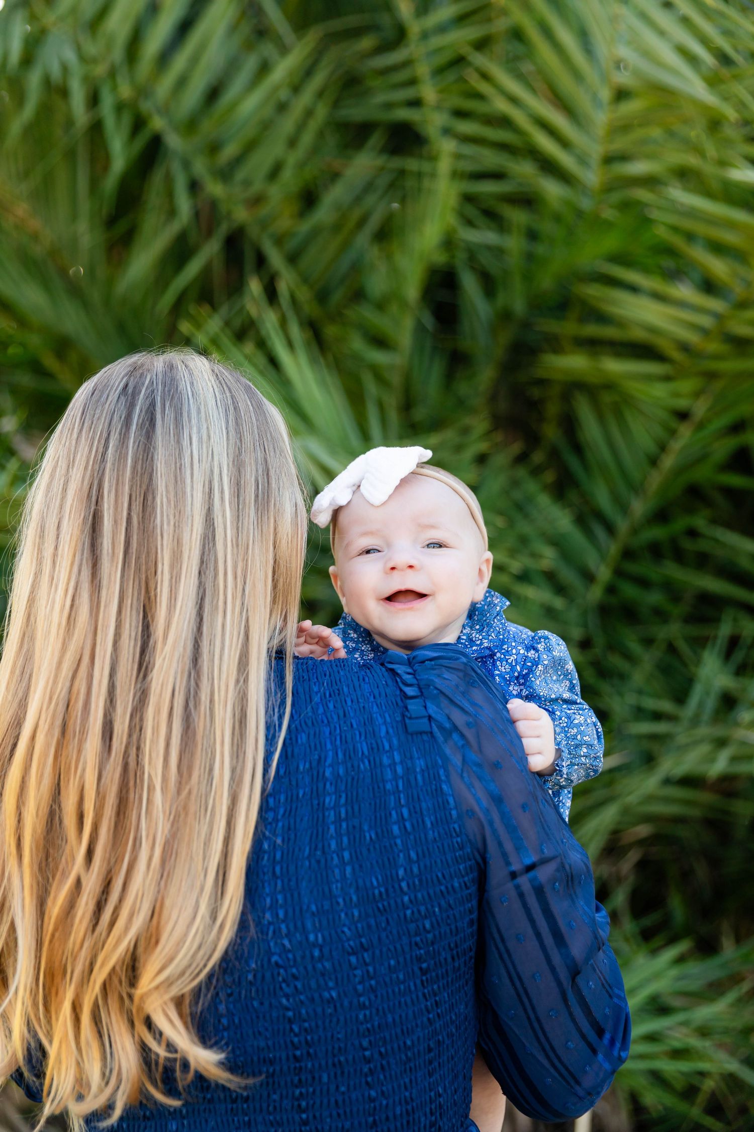 Blonde mother holds smiling baby girl with white bow headband against lush green palm backdrop at the University of Tampa.