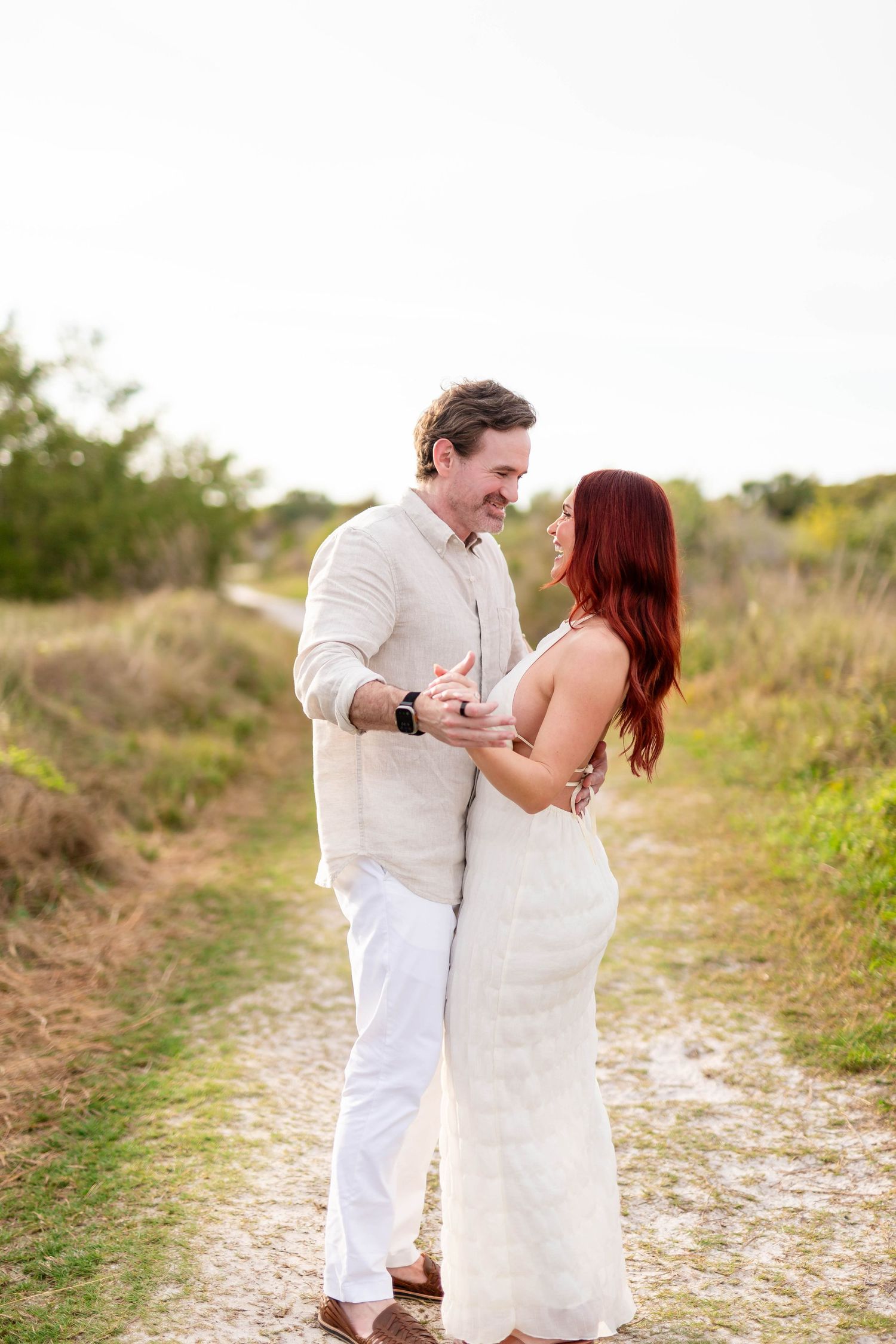 Couple laughing and embracing on a sandy trail surrounded by green shrubs during golden hour light at Honeymoon Island.
