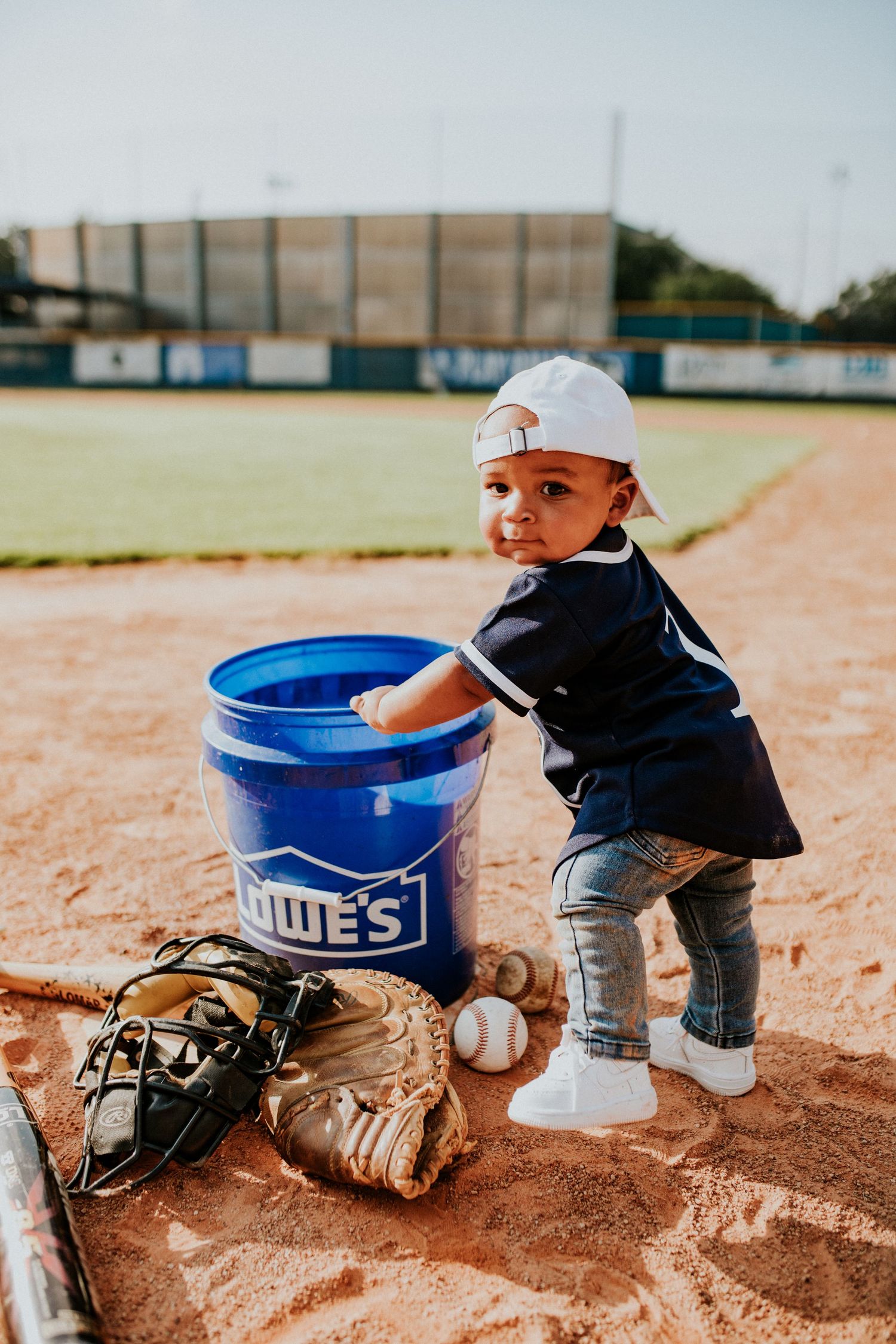 Adorable toddler in baseball outfit stands beside a Lowes bucket on a baseball field.
