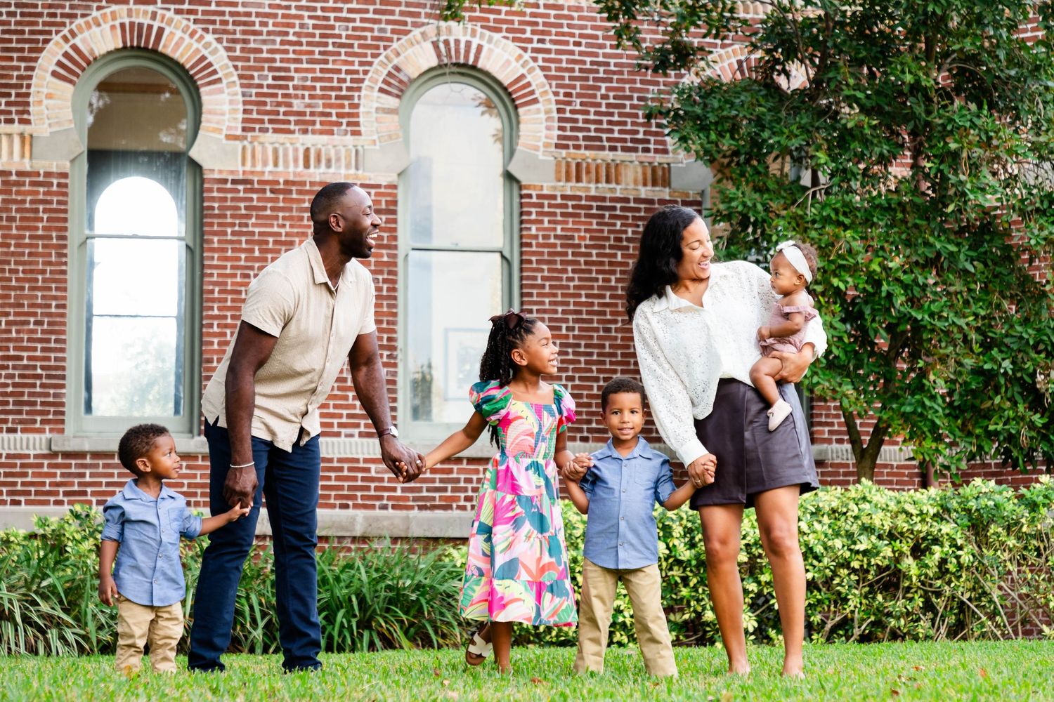 Joyful Black family with four young children standing on grass in front of a brick building at the University of Tampa.