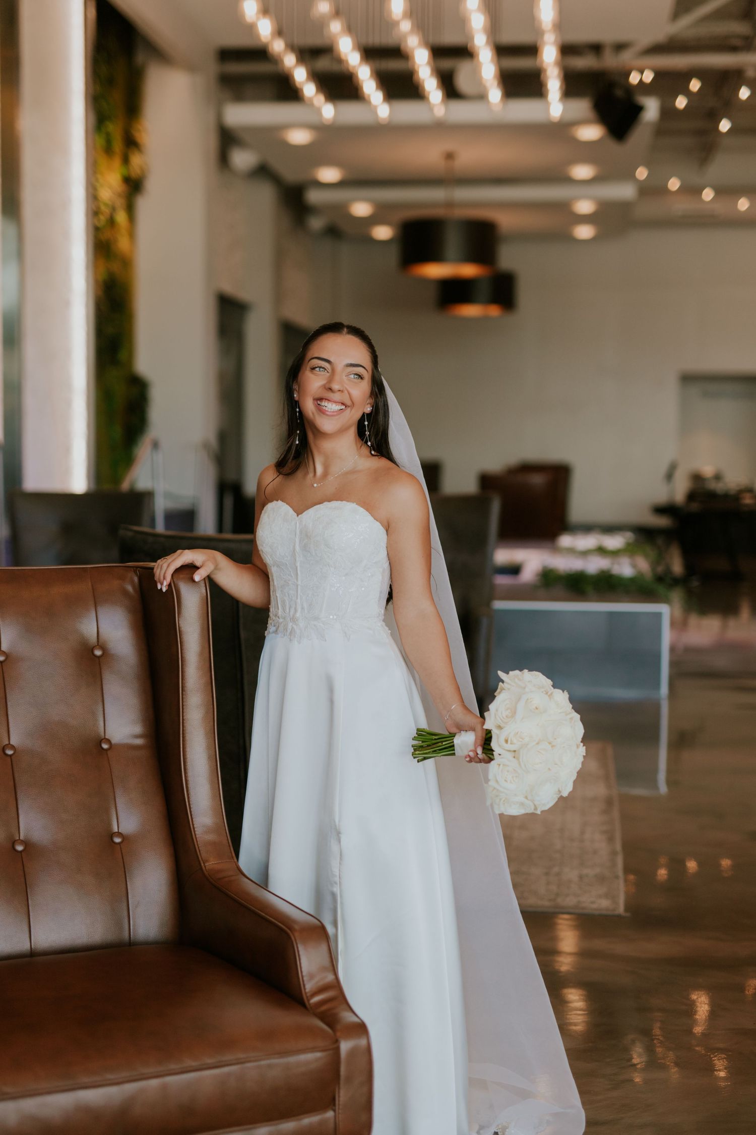 Smiling bride in strapless white gown holds white peony bouquet in a modern wedding venue interior.