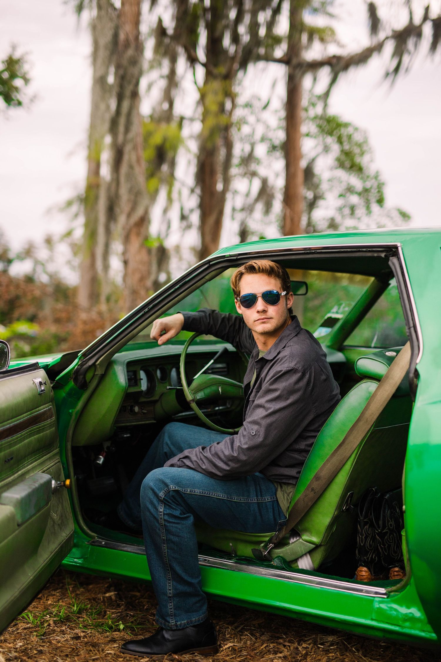 Young man in sunglasses sitting in open door of vintage green muscle car in wooded area.